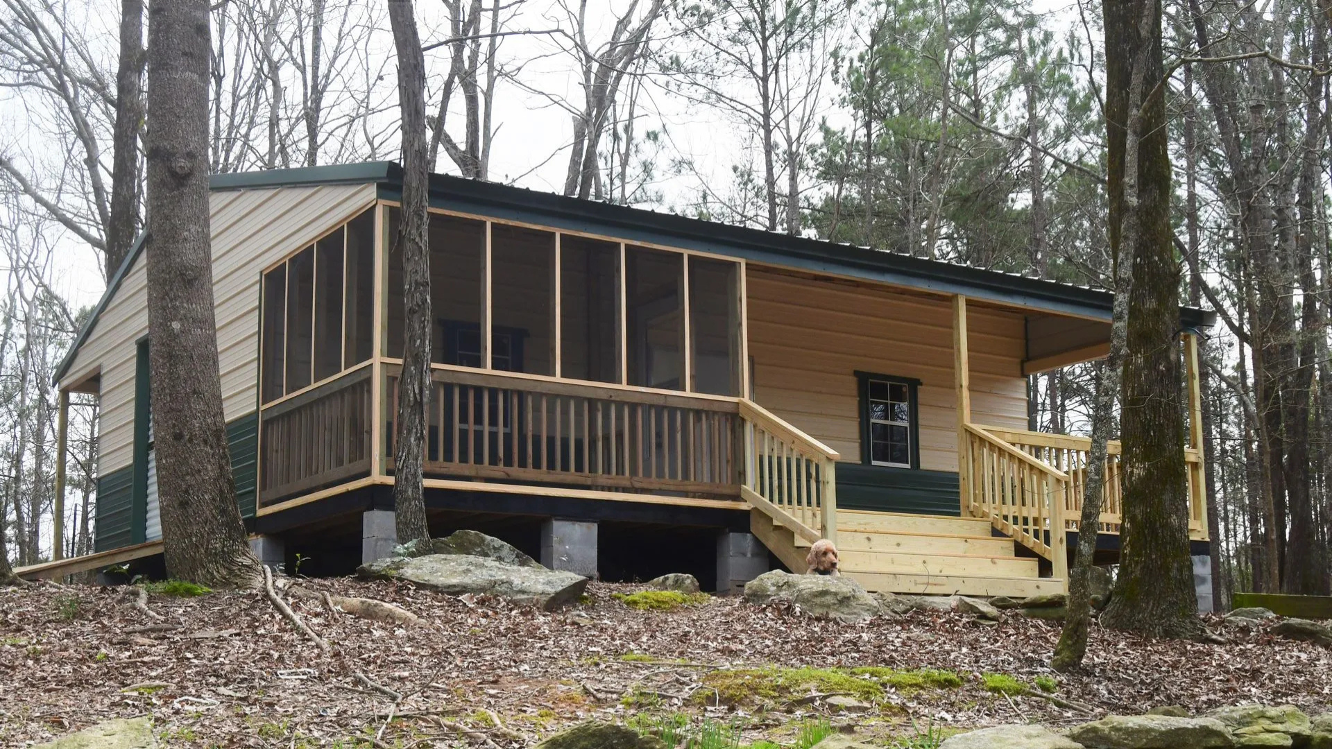 Cabin with a screened porch, wooden deck, and dark green roof, in a wooded area.