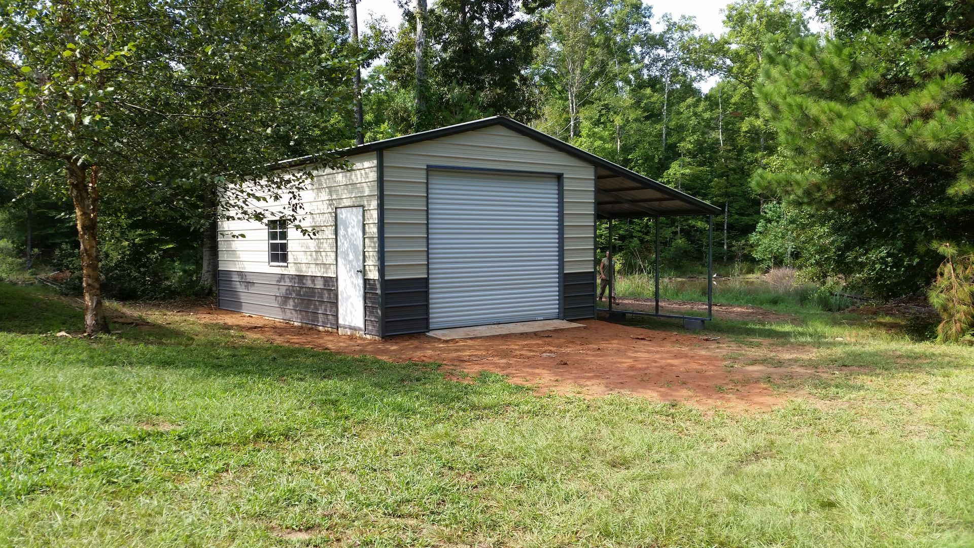 Metal shed with garage door and lean-to in a grassy yard, surrounded by trees.