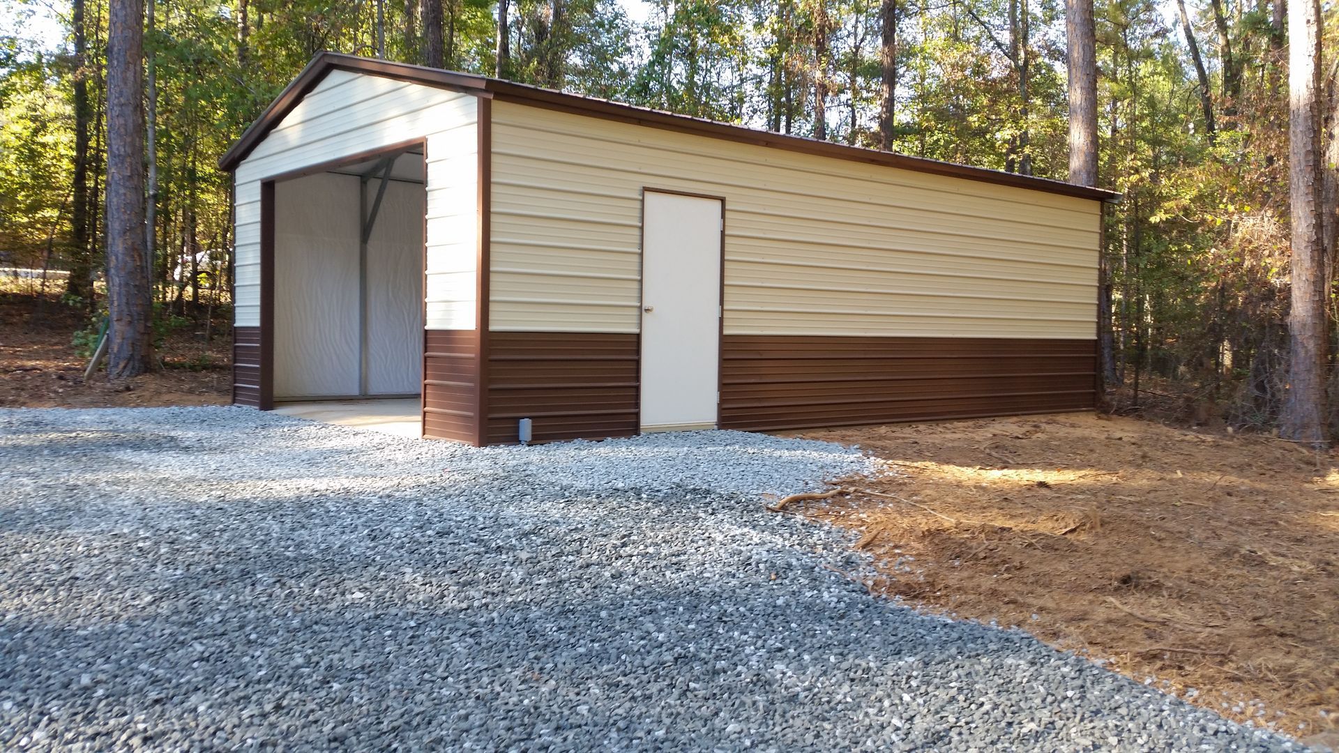 Tan and brown metal garage with open bay door and gravel driveway, surrounded by trees.