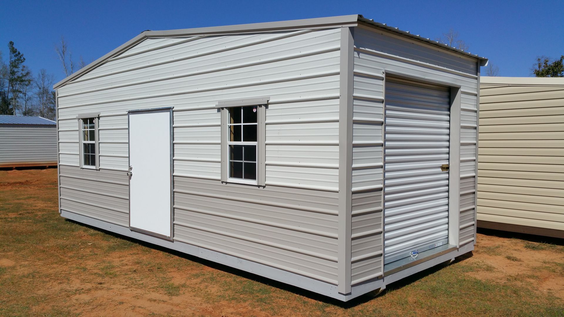 A white metal shed with a garage door is sitting in the middle of a field.