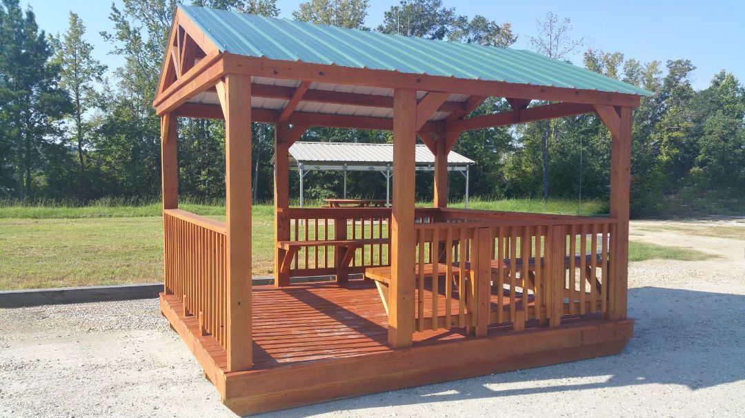 A wooden gazebo with a green roof is sitting in a gravel lot.