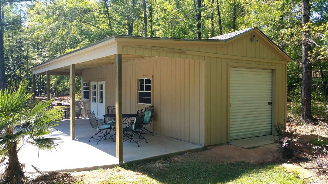 Tan shed with a concrete patio, seating area, and a garage door in a wooded area.
