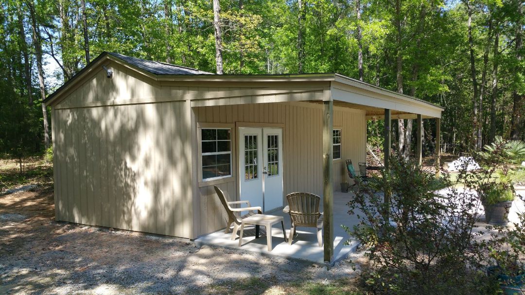 Beige shed with porch, two doors, two chairs, and surrounded by trees.
