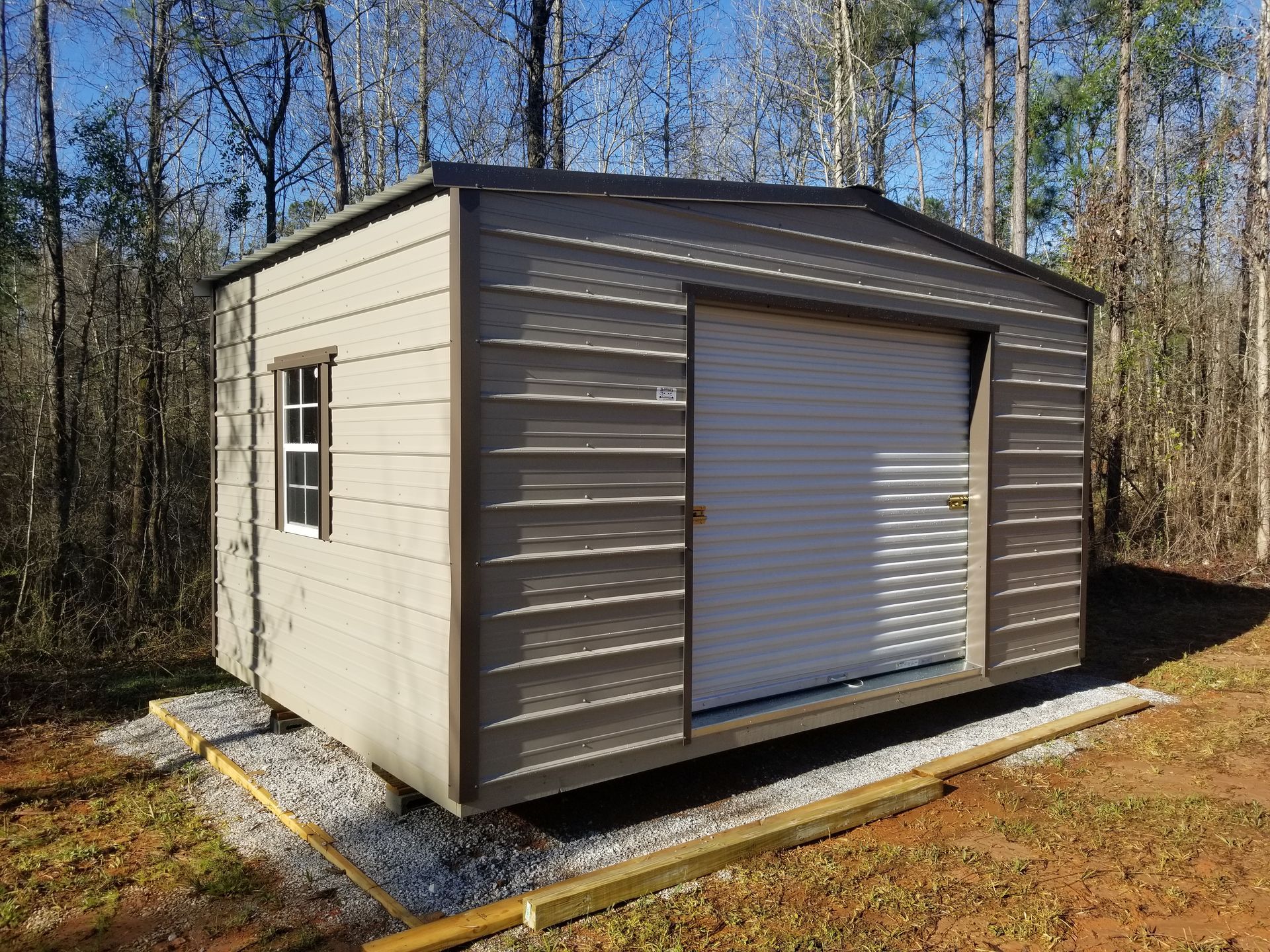 Tan shed with roll-up door and small window, set on gravel base in a wooded area.