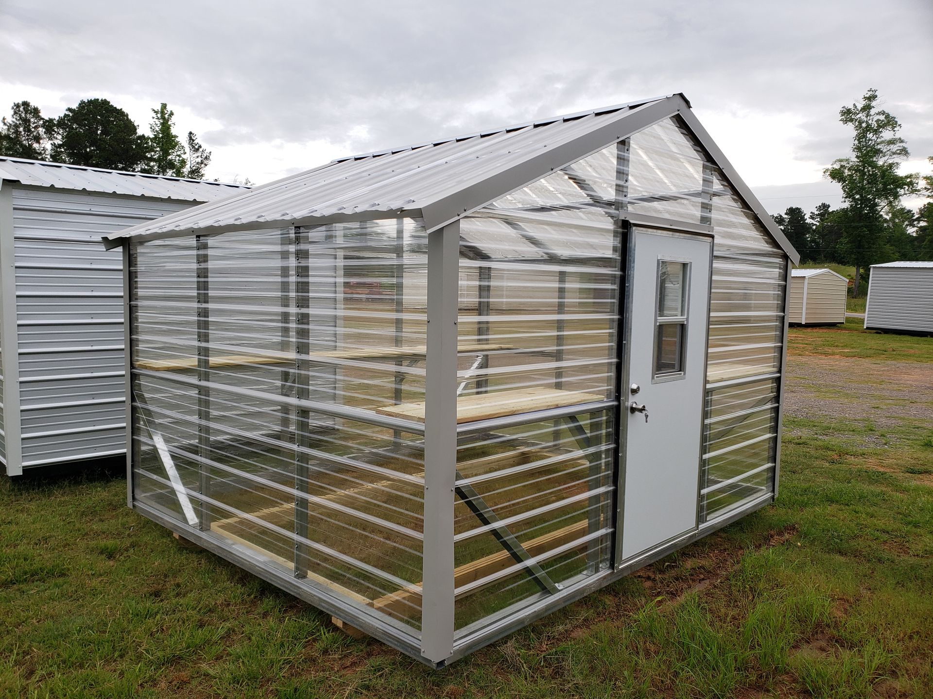 A small, clear greenhouse with a white door and metal frame sits on grass under a cloudy sky.