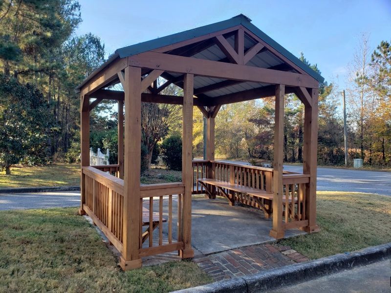 Brown wooden gazebo with benches, on a concrete pad, surrounded by grass and trees.