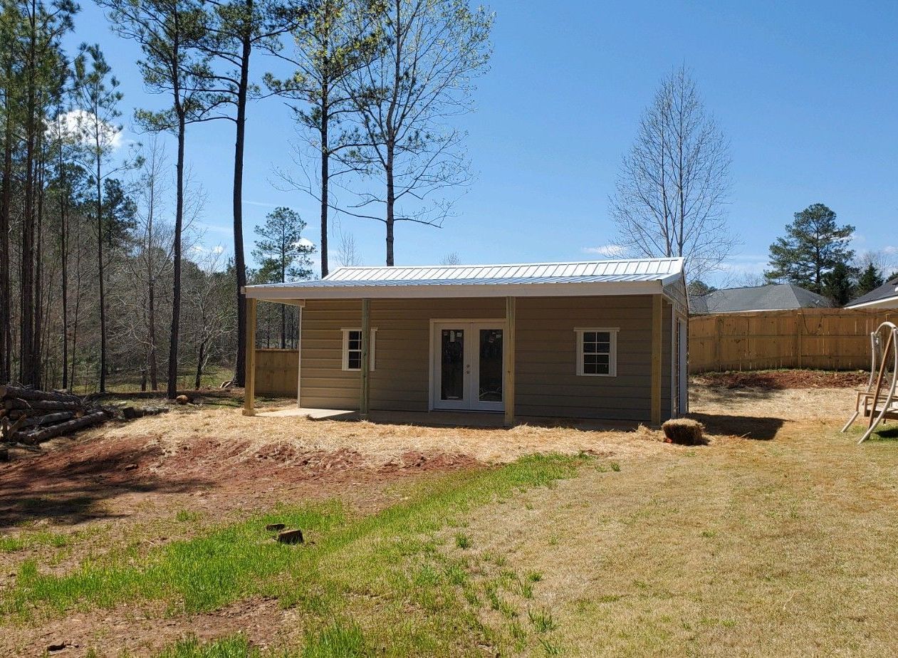 Small beige building with white roof, set in a yard with trees and a wooden fence.