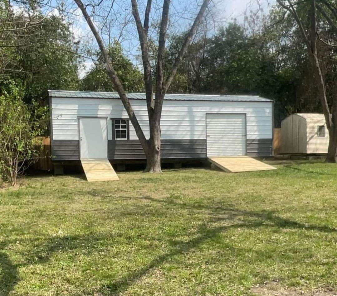 Metal shed with a door and garage door. Ramps lead to each. A small window and a tree are also present.