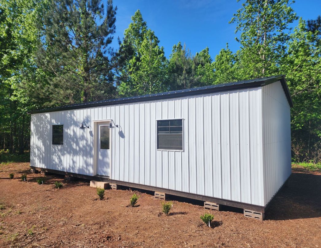 White metal-sided rectangular building with a dark roof, two windows, and a door; set in a wooded area.