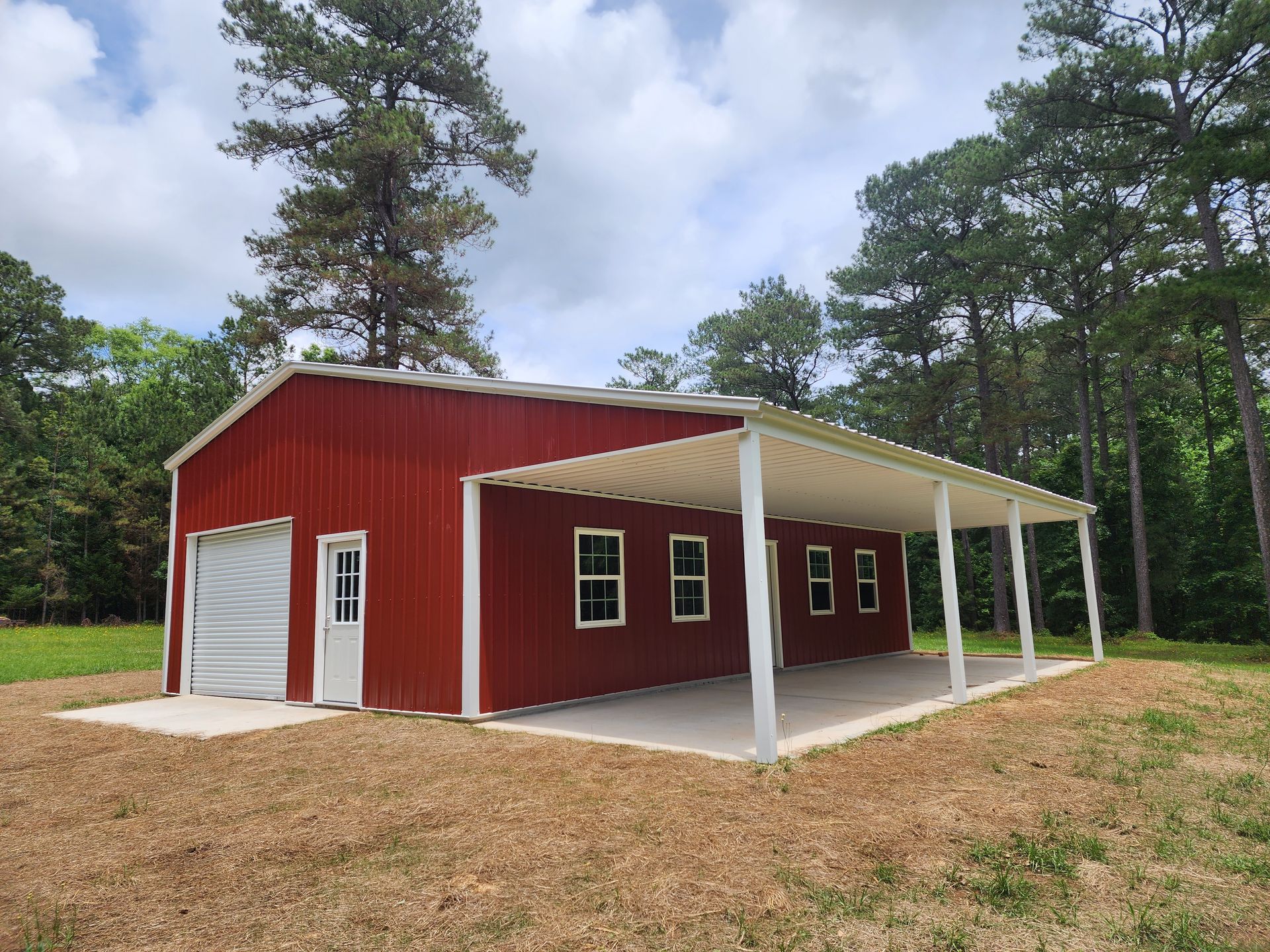 Red metal building with white trim, garage door, and covered porch on a concrete slab in a wooded area.