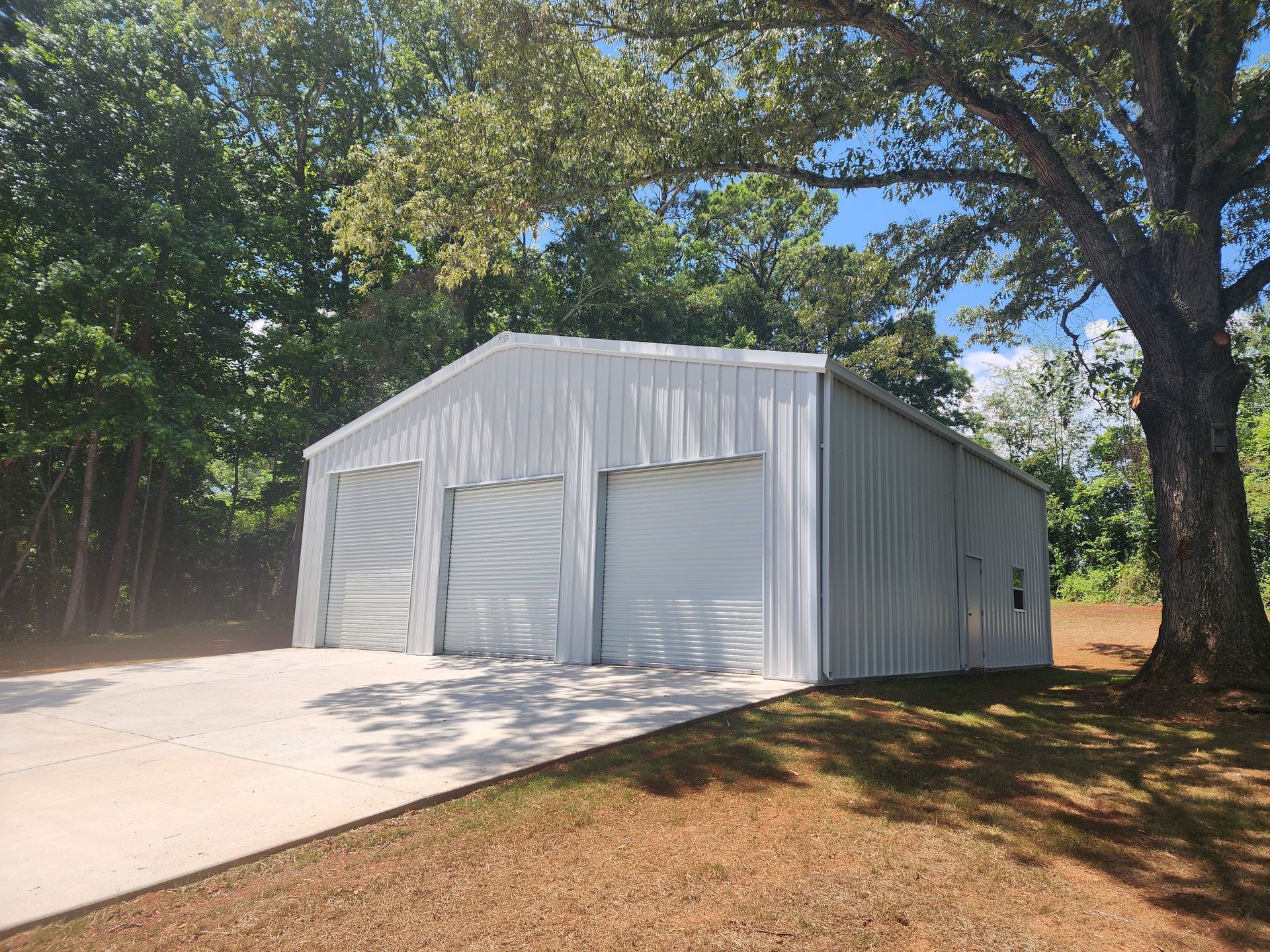 A white metal garage with three garage doors is surrounded by trees.