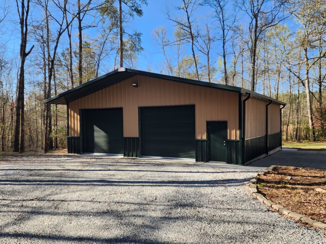Tan and black metal building with two garage doors, a small door, and a gravel driveway, set in a wooded area.