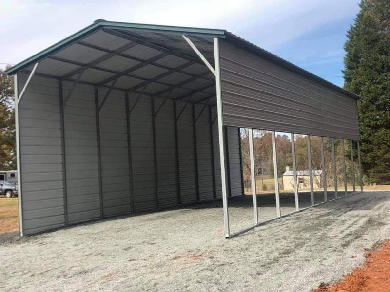 Gray metal carport on a gravel surface. Green roof, open sides, and a clear sky background.