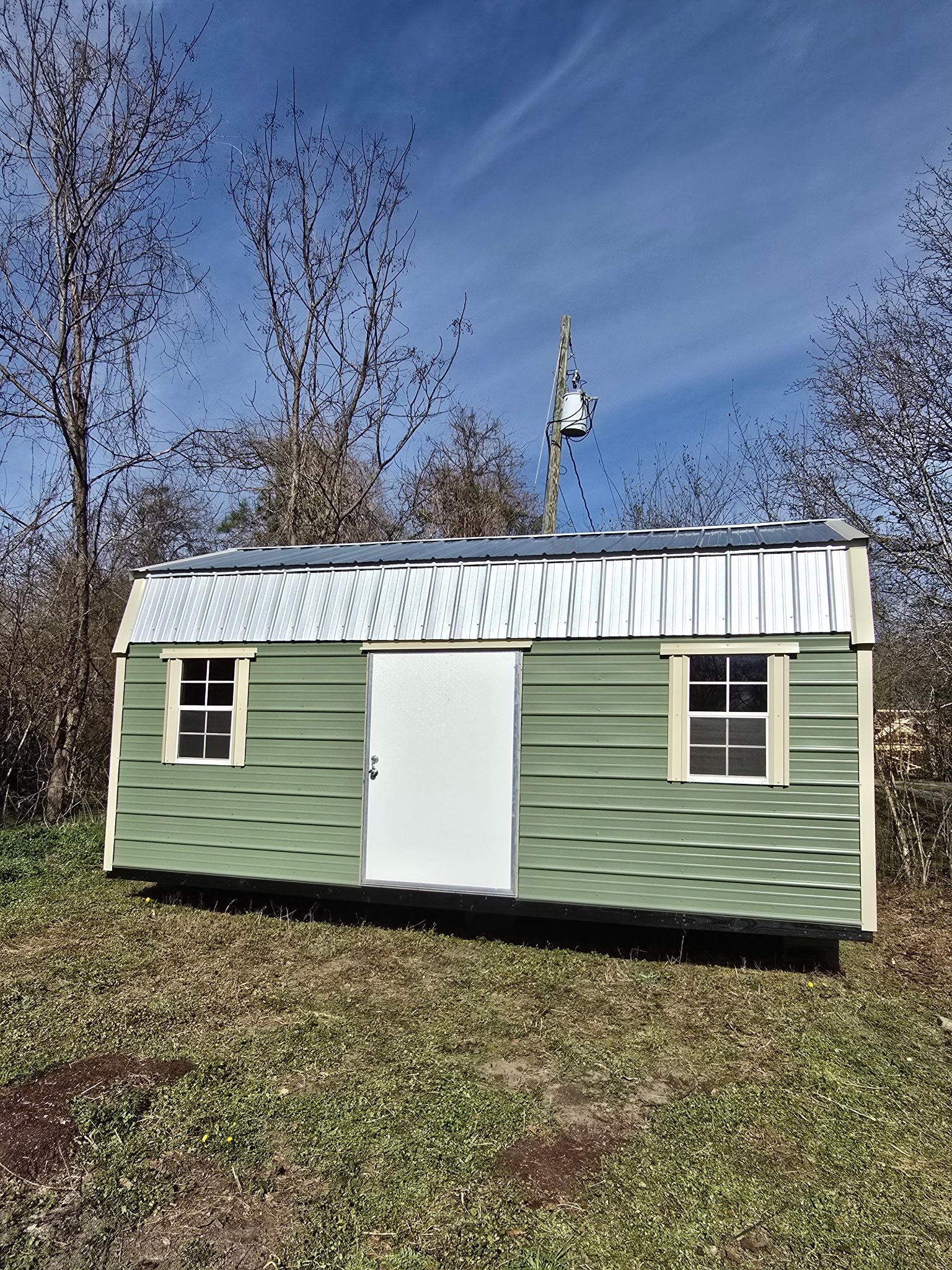 Green and white shed with a metal roof on a grassy field, with trees and blue sky.