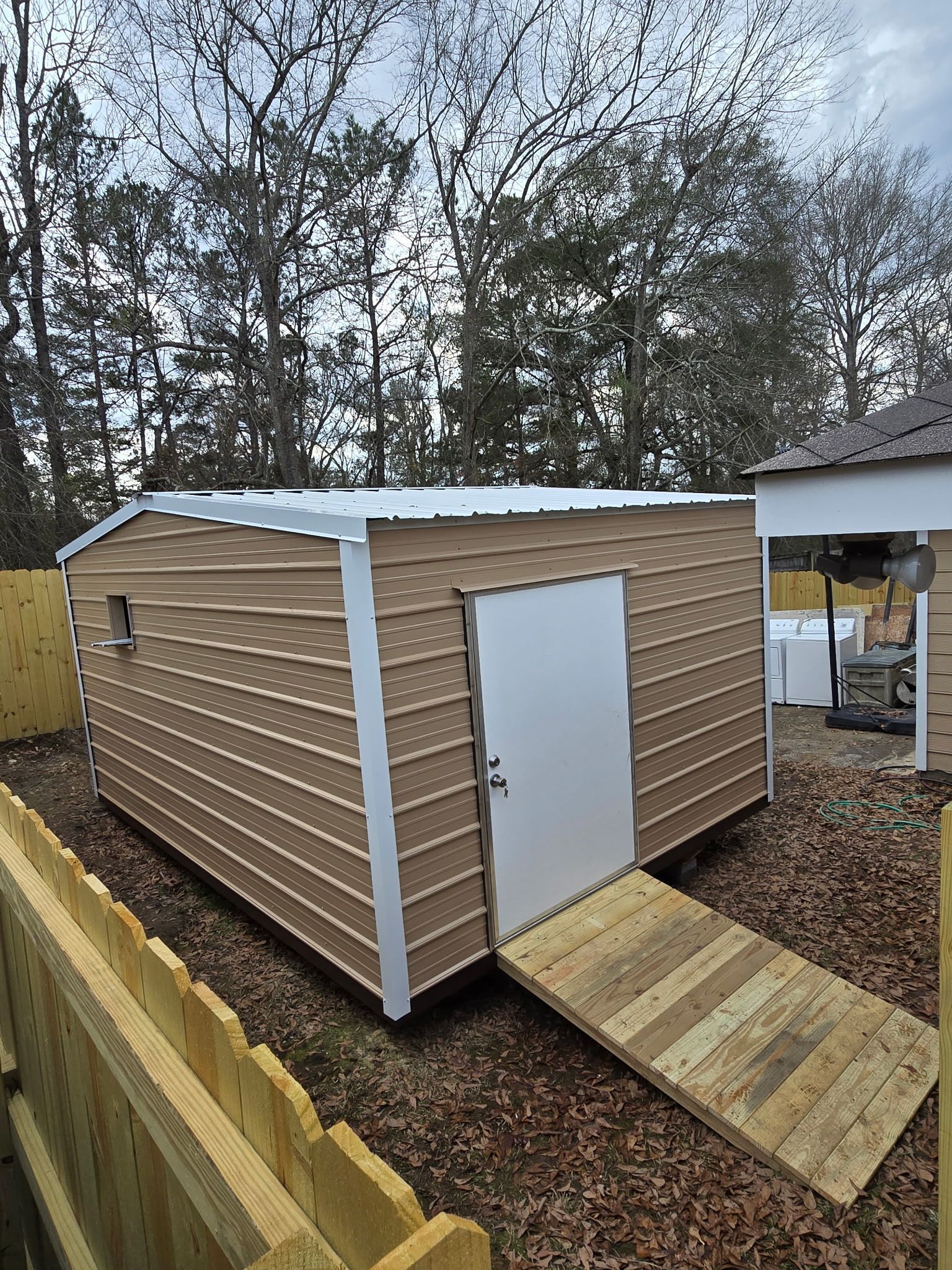 Tan shed with a white door and ramp. It is surrounded by a wooden fence and trees.