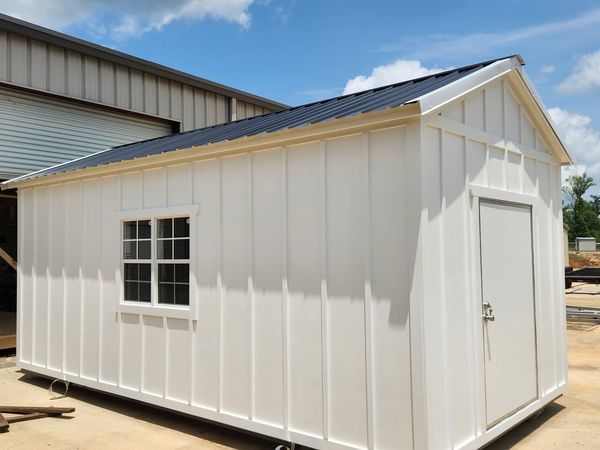 White storage shed with black metal roof, window, and door against a blue sky.
