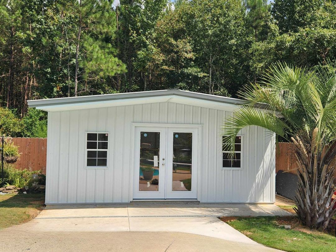 White shed with double doors and two windows, set in a yard with a palm tree and trees in the background.