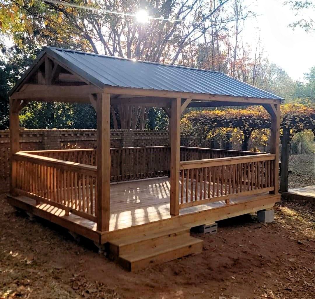 A wooden gazebo with a metal roof is sitting in the middle of a forest.