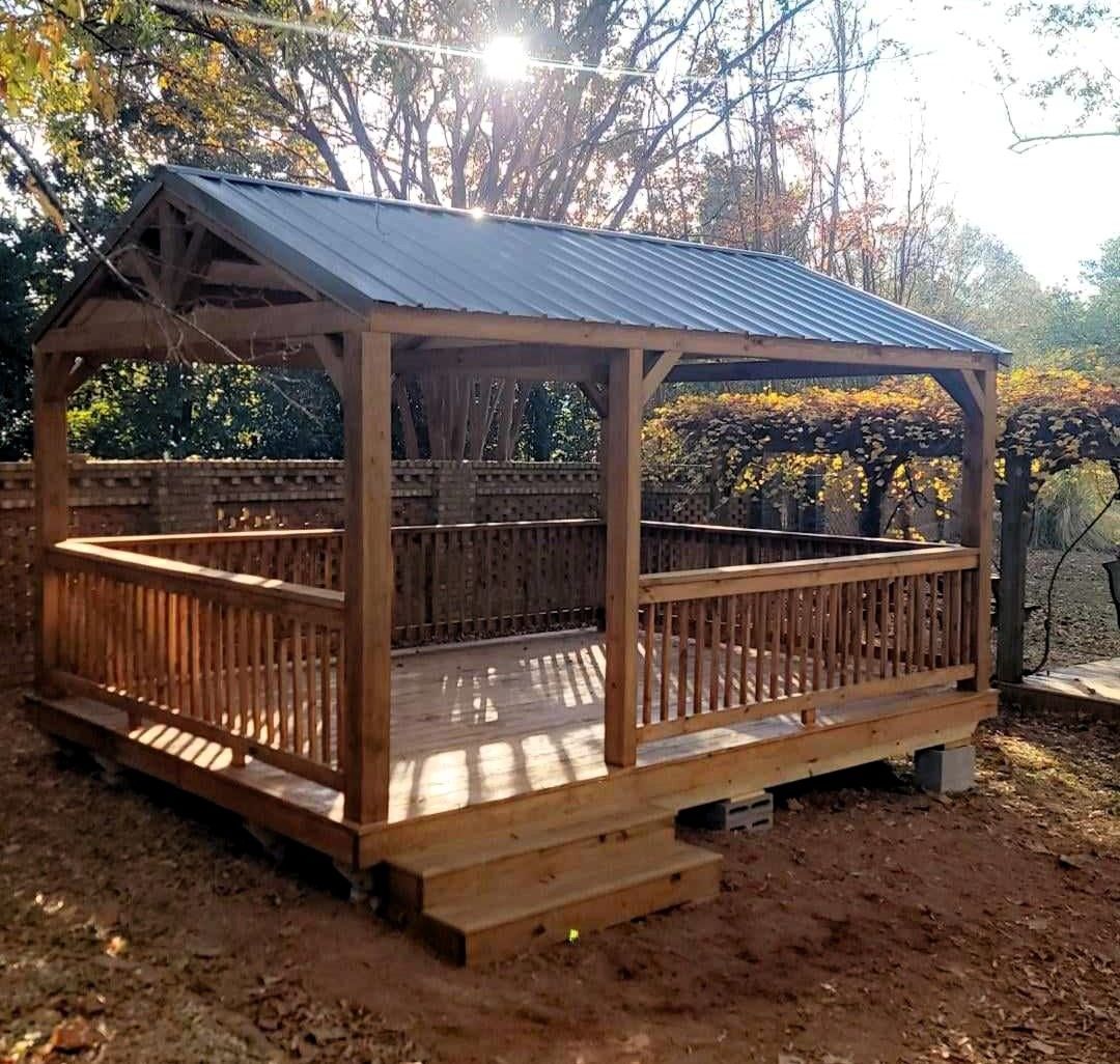 Wooden gazebo with metal roof, railing, and steps, in a yard, with sunlight.
