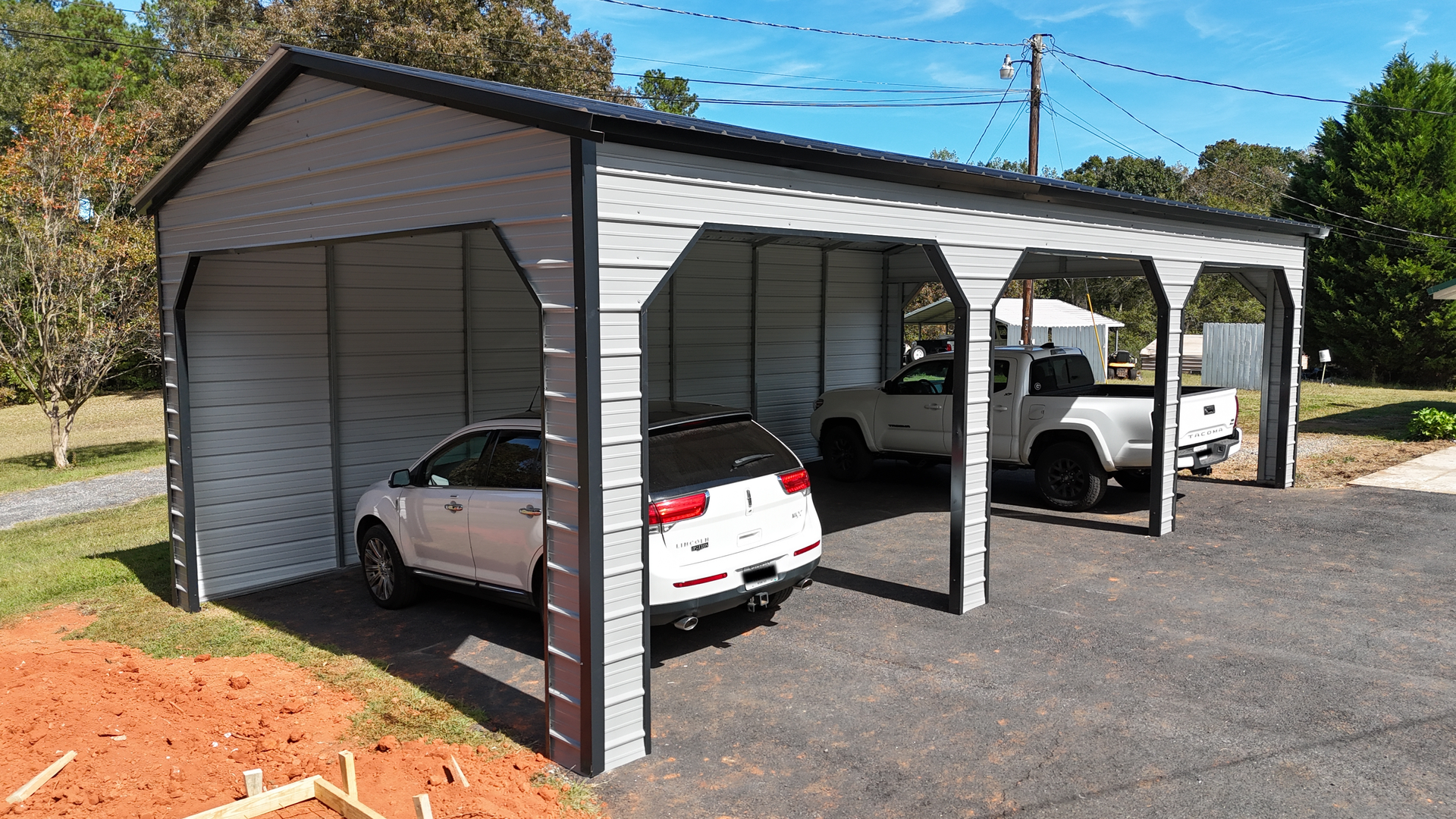 Gray carport sheltering white vehicles on a paved surface, set against a green lawn and trees.