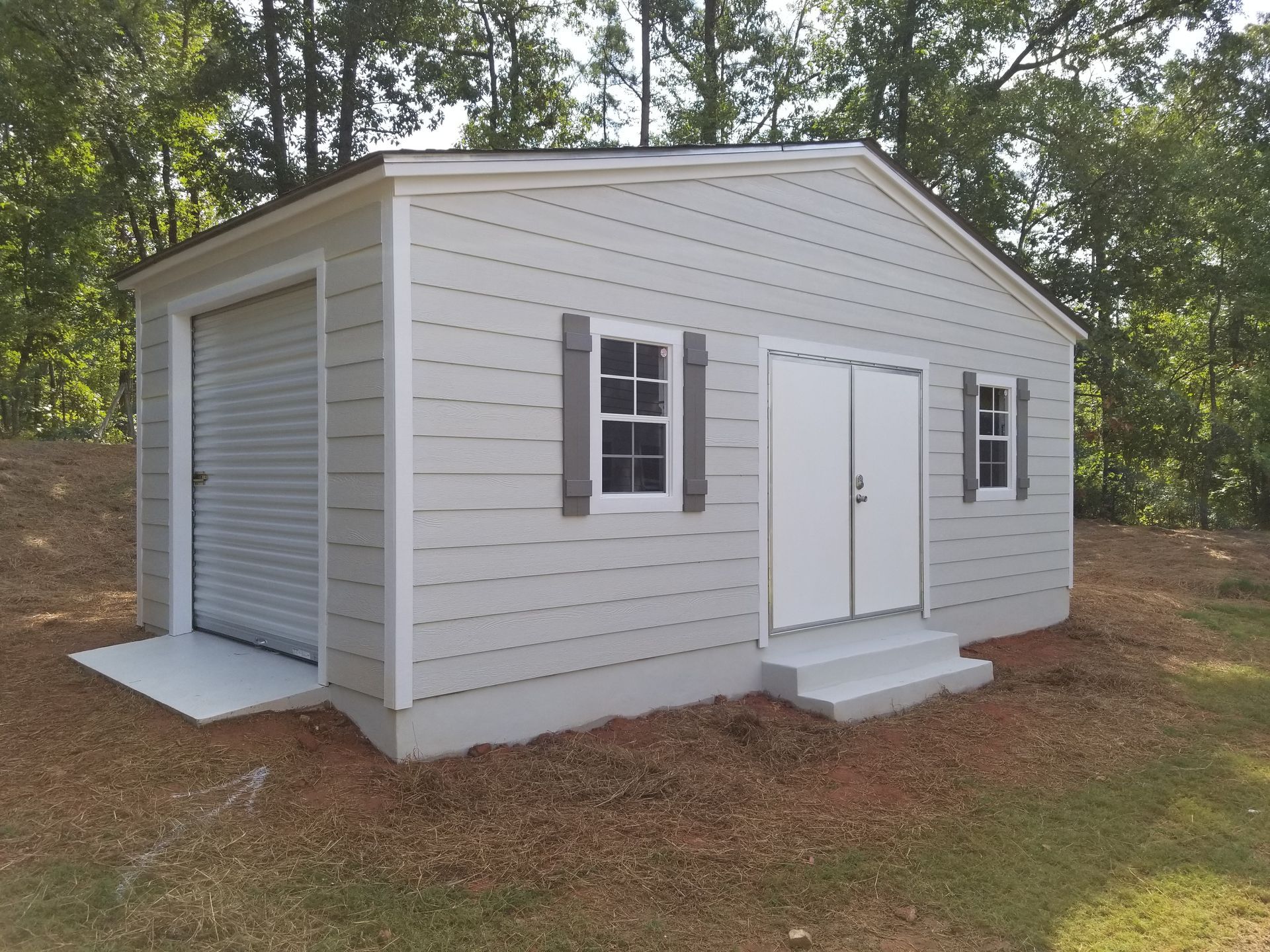 Light gray shed with a roll-up door, double doors, two windows with shutters, and a concrete ramp/steps.