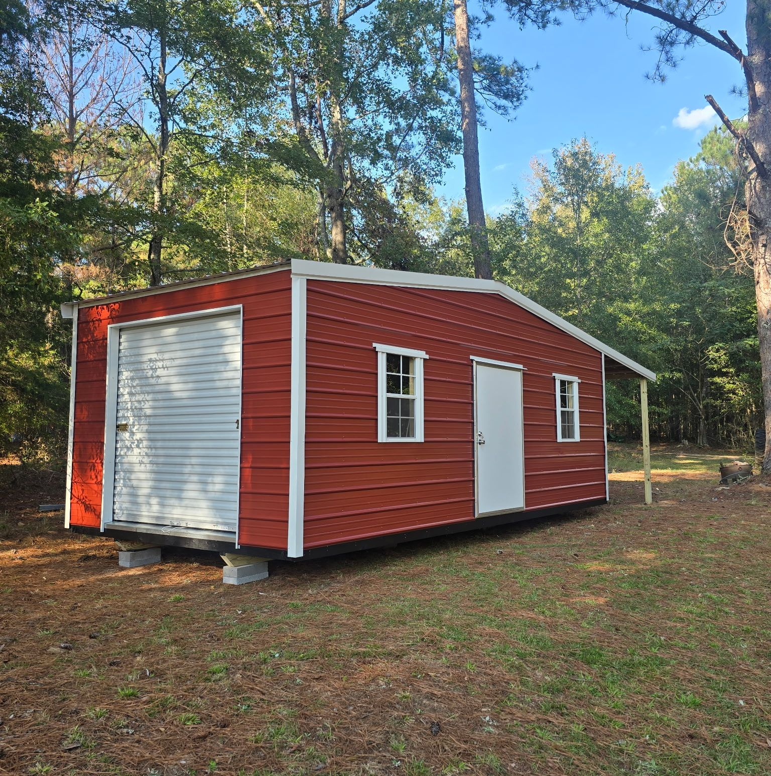 Red shed with white door, garage door, and windows in a wooded area.