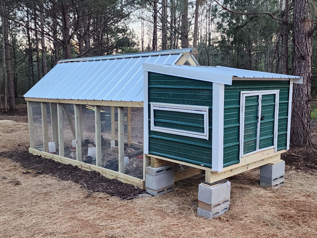 A green chicken coop with a metal roof is sitting in the middle of a field.