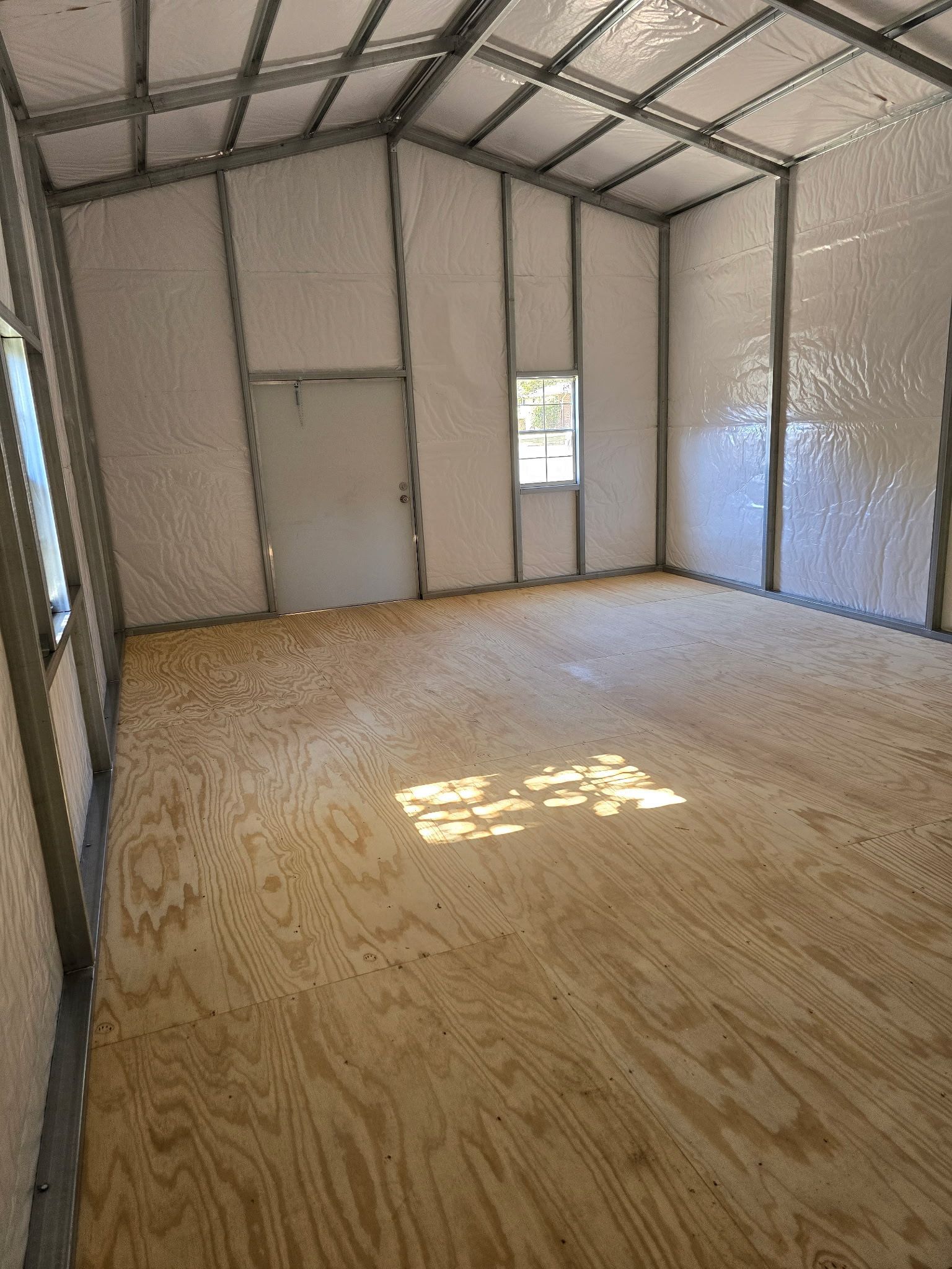 Interior of a metal frame building with plywood floor, door, window, and insulated walls.