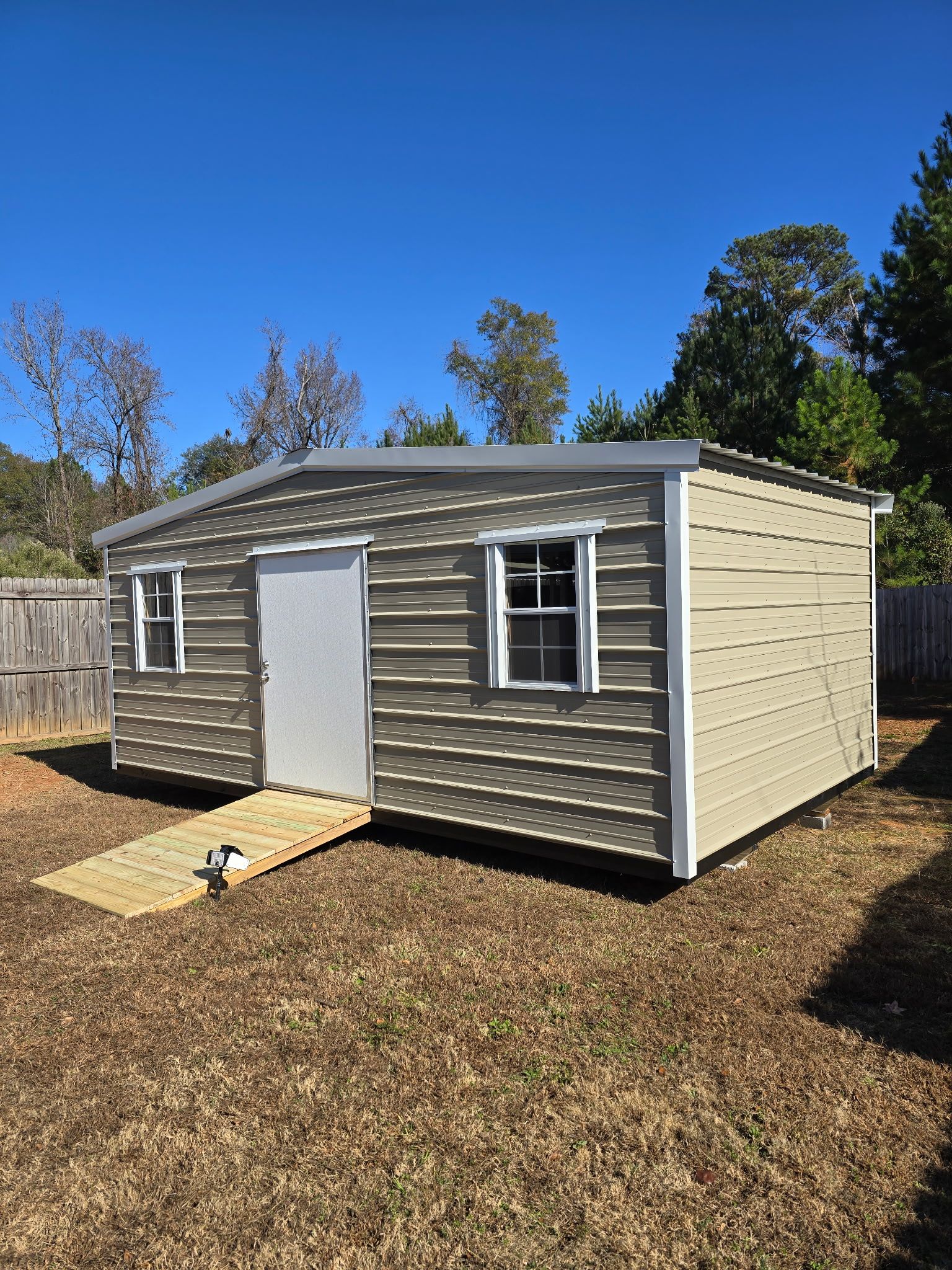 Tan and white metal shed with a ramp, two windows, and door, set in a yard.