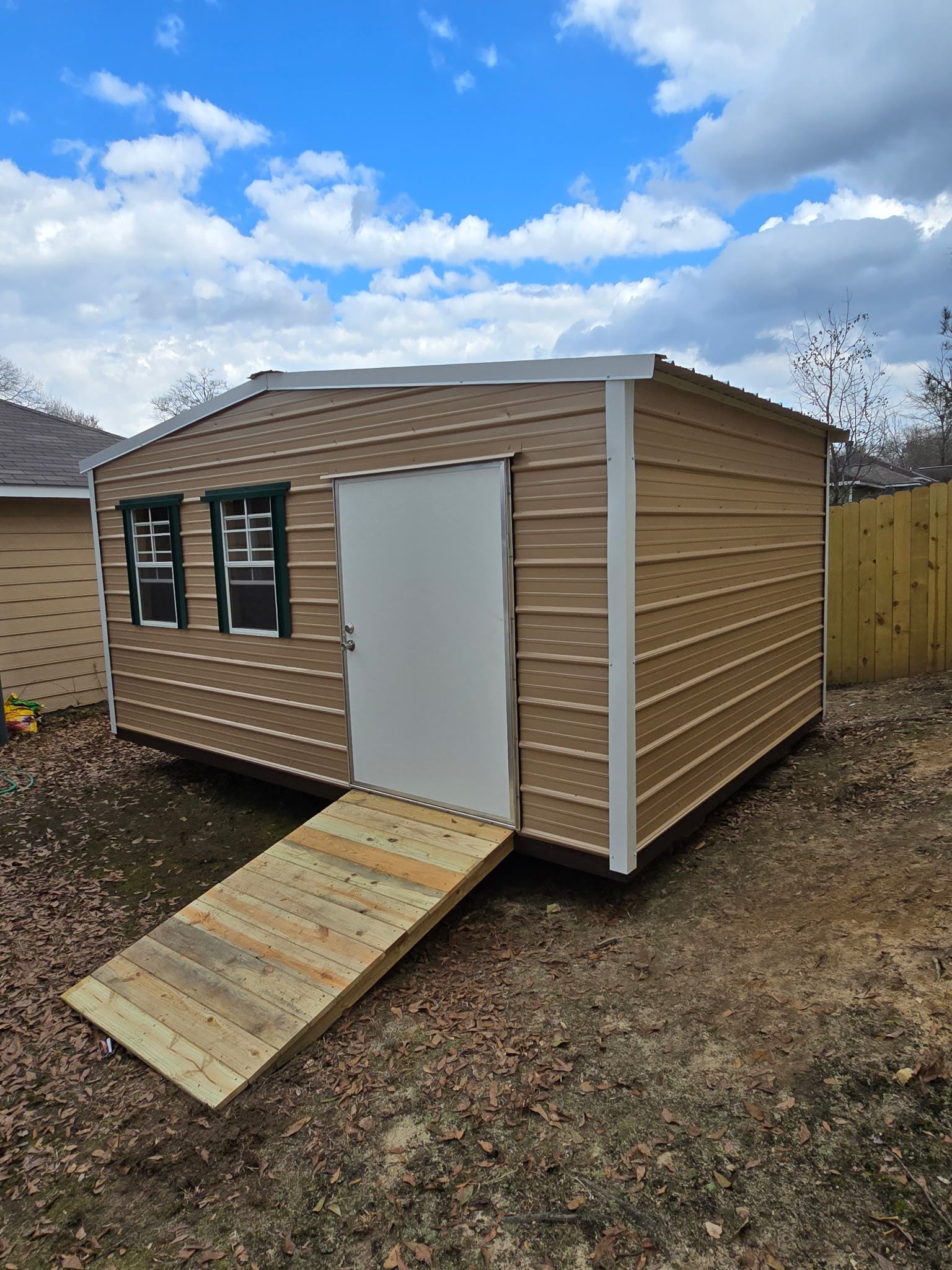 Tan shed with white door, ramp, and green-trimmed windows under a cloudy blue sky.