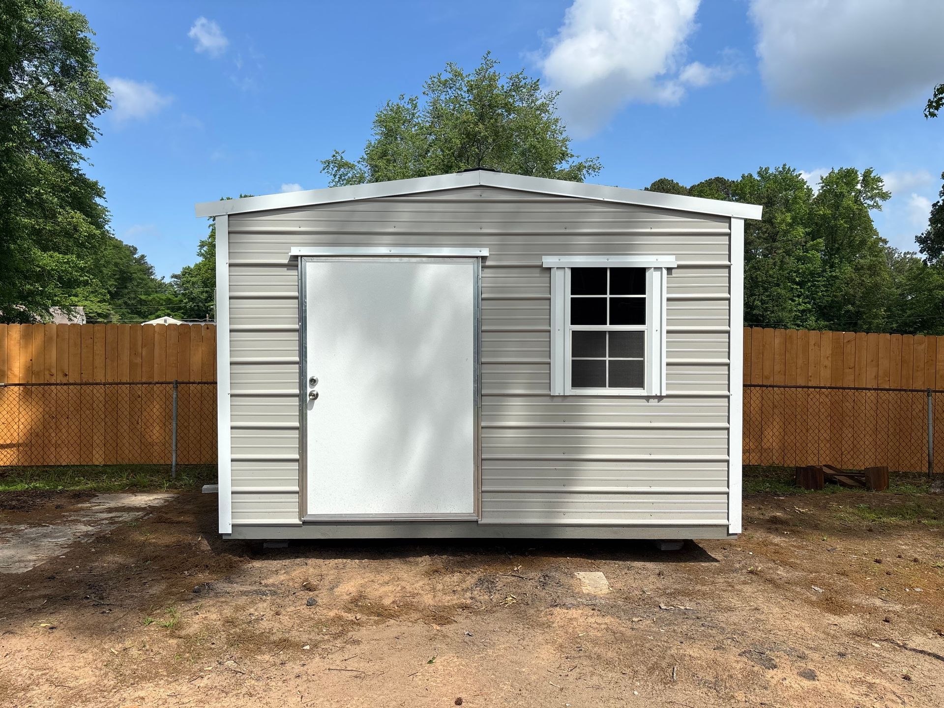 Gray metal shed with a door and a window, set against a wooden fence and blue sky.