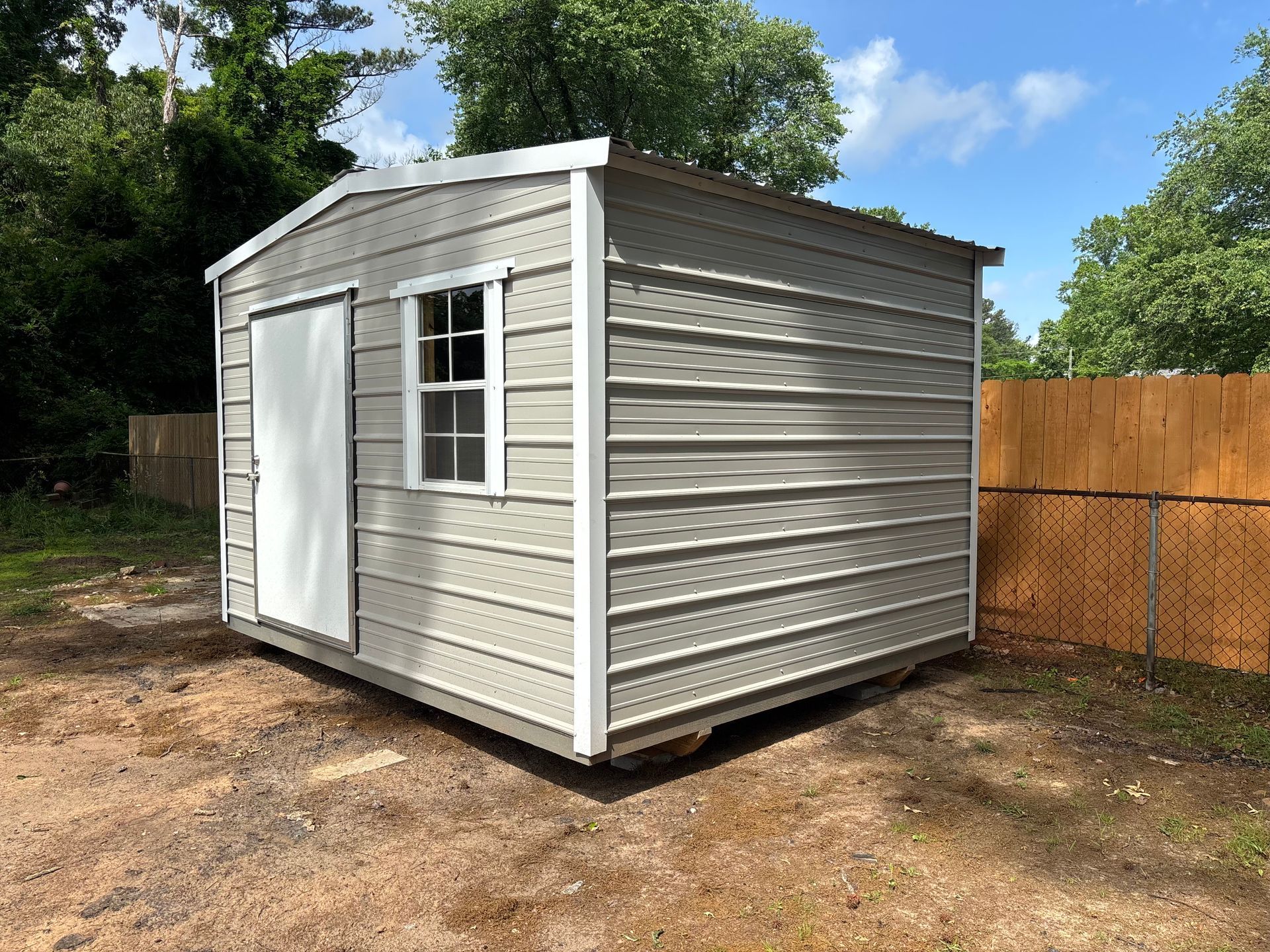 Gray metal storage shed with white door and window, set in a yard.