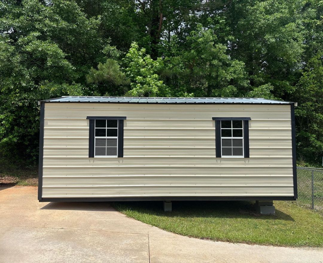 A small white metal building with two windows sitting on grass and pavement