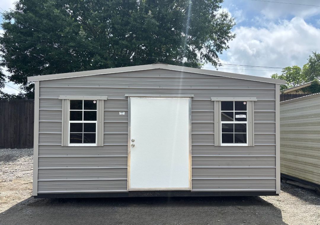 A small shed with a white door and windows is sitting in a gravel lot.