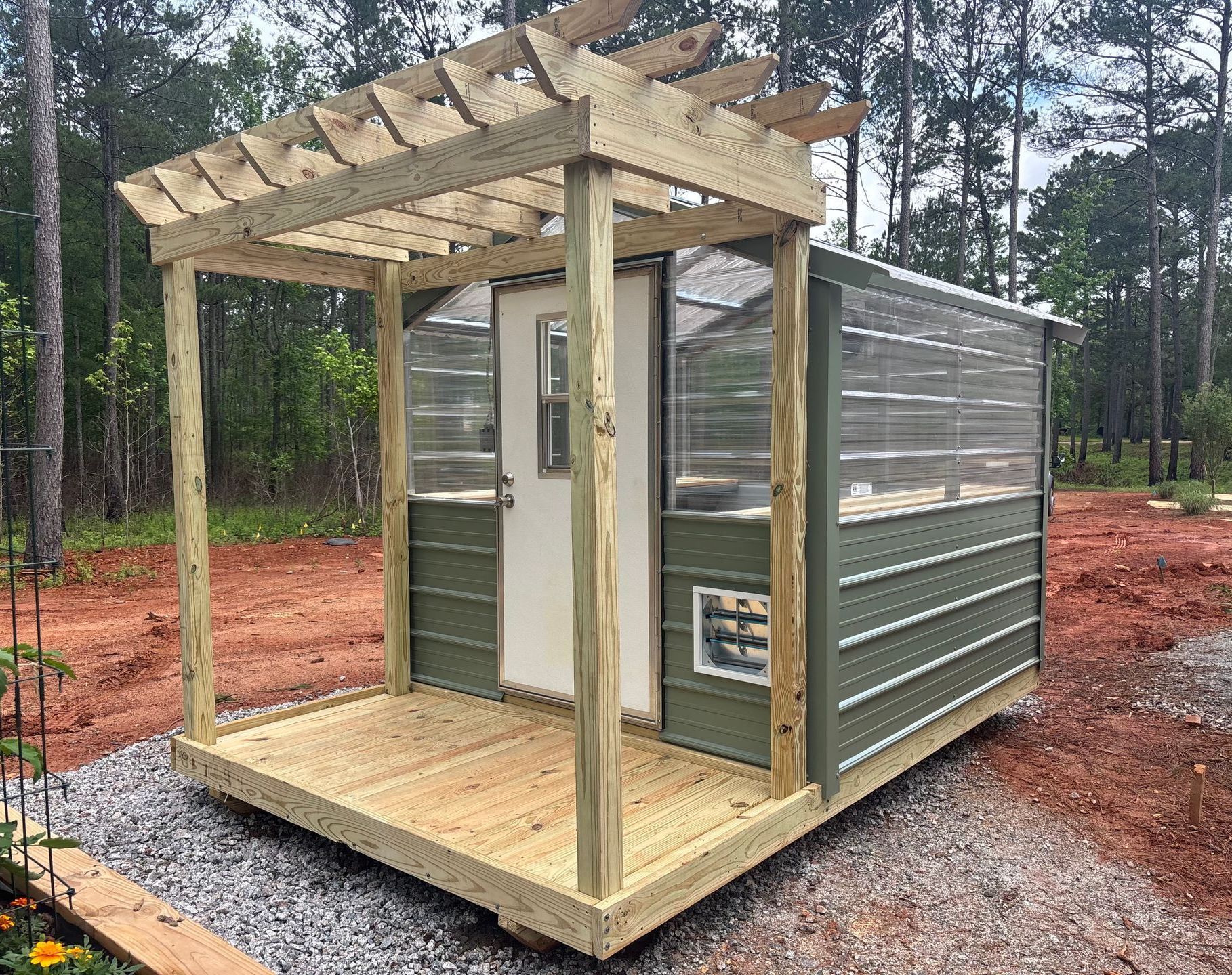 A small greenhouse with wooden deck and pergola, painted green and tan, in an outdoor setting.