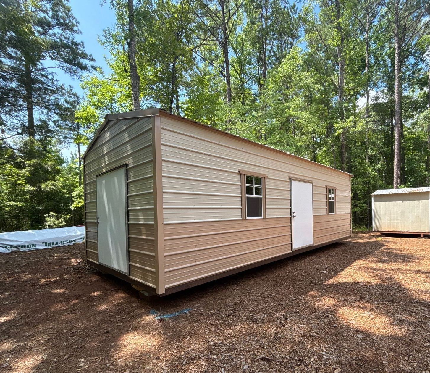 Tan and cream-colored storage shed with two doors and a window in a wooded area on a sunny day.