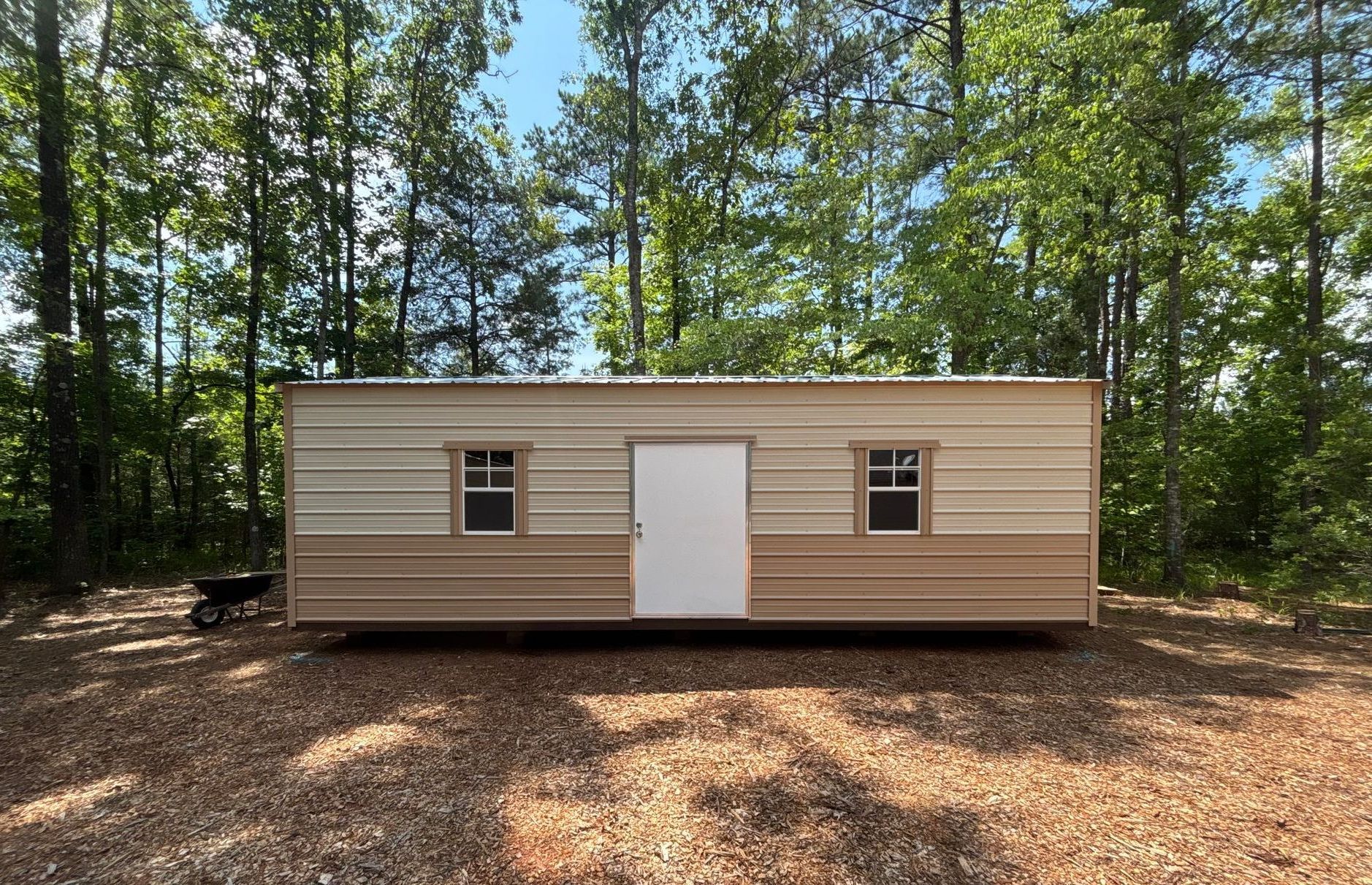 Tan and beige metal shed with a white door and two windows, set in a wooded area.