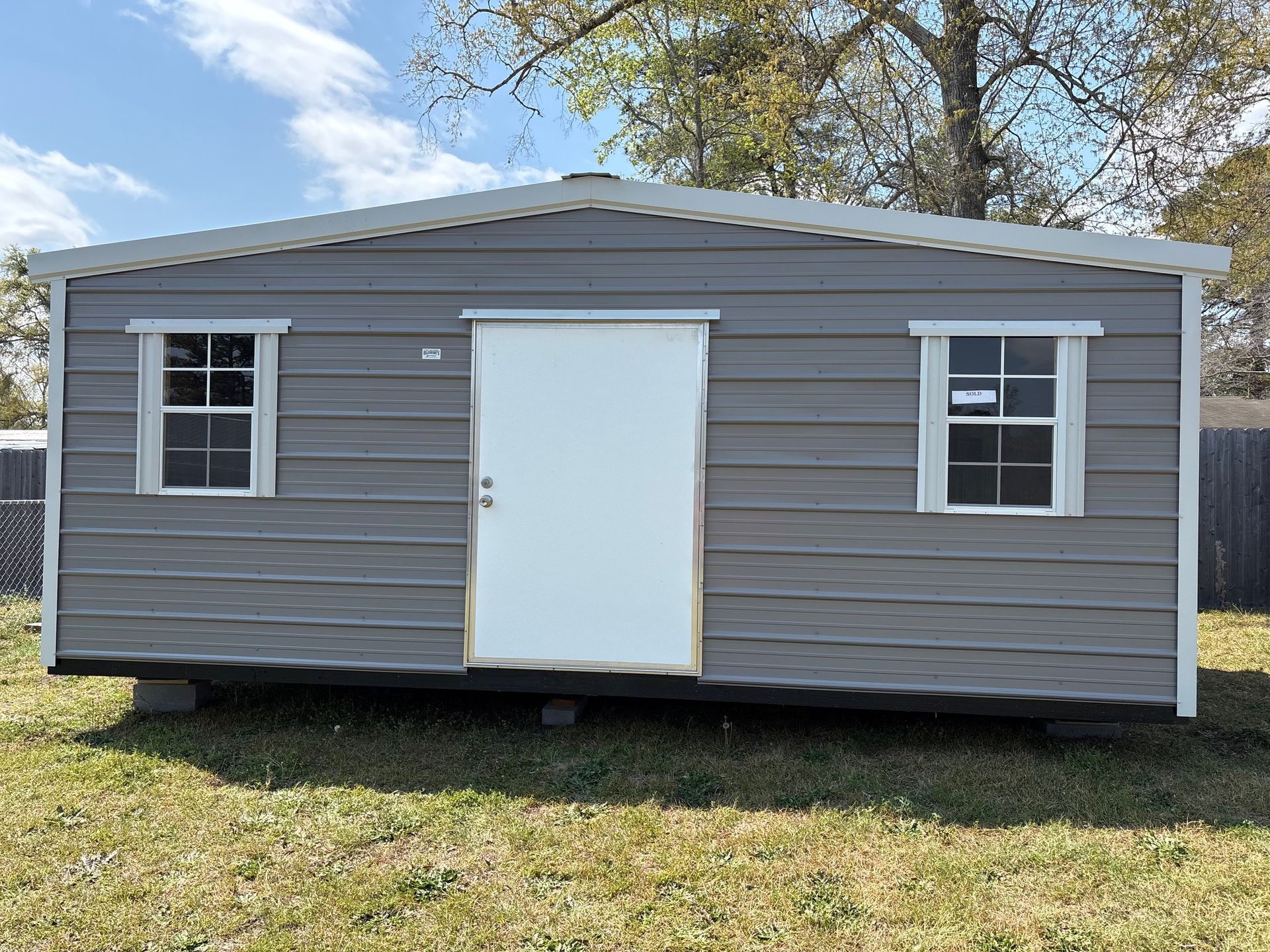 A gray, rectangular storage shed with a white central door and a single window on each side, sitting on a grassy lot.