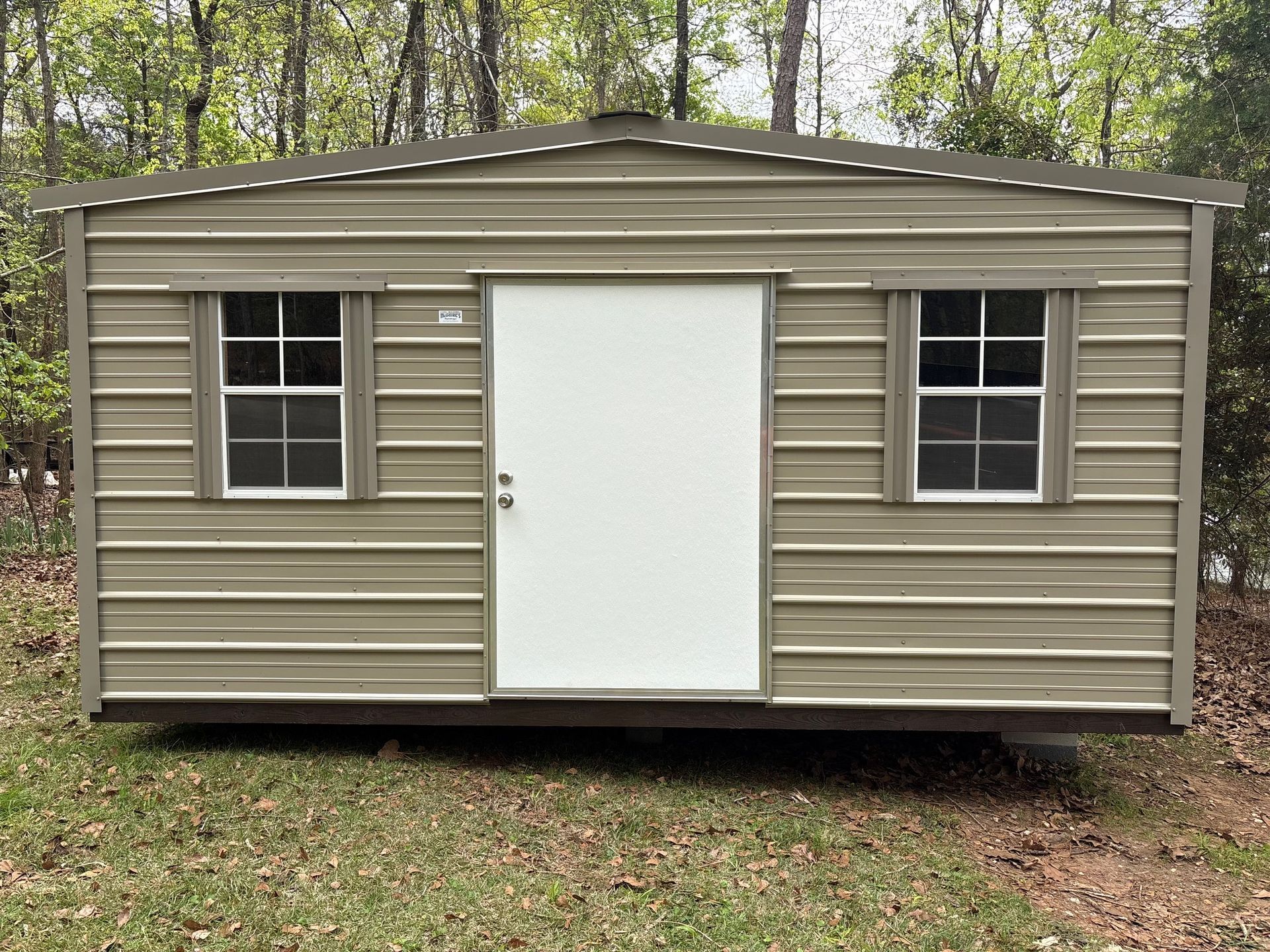 Tan metal storage shed with two windows and a central white door, set on grass in a wooded area.