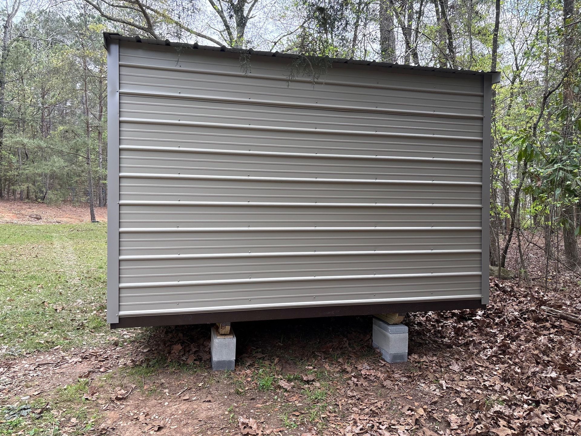 A rectangular, tan metal shed with brown trim sitting on cinder block piers in a wooded, grassy area.