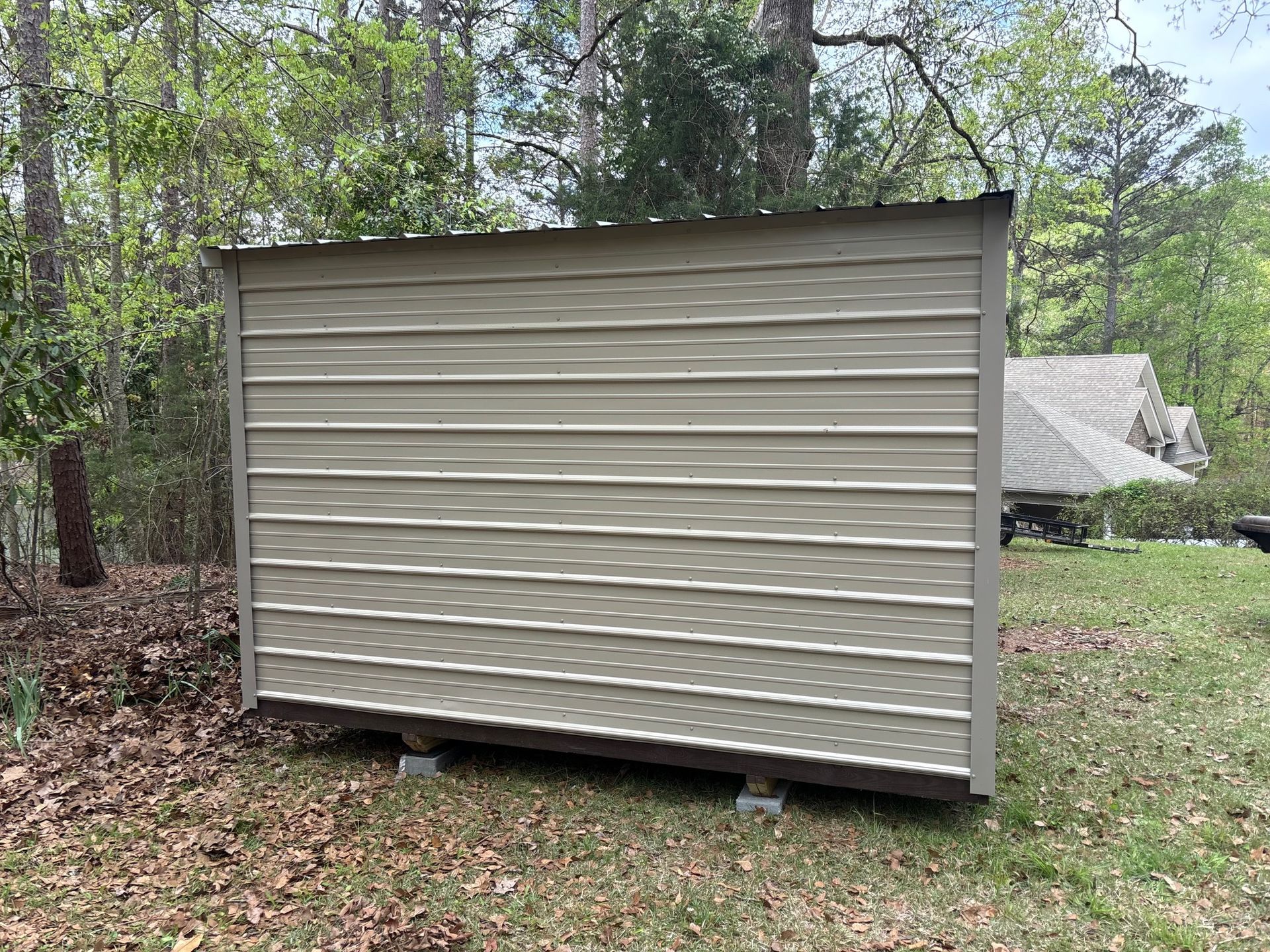 A beige metal storage shed sits on a gravel base in a grassy, wooded yard.