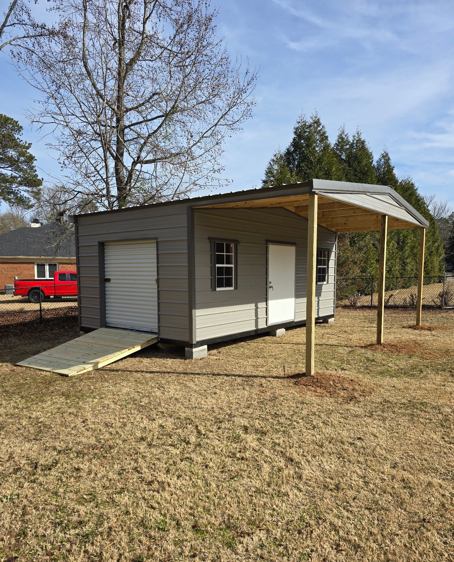 Gray shed with a ramp, white door, and carport, on a grassy lot.