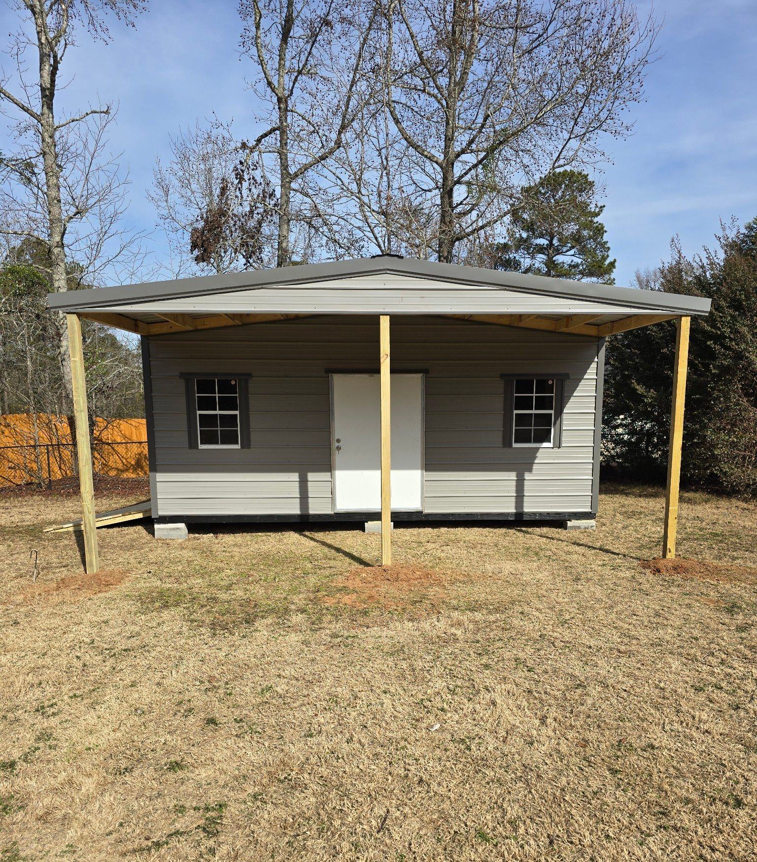 Gray shed with metal roof and awning, two windows, door, and two support posts on a grassy lot.