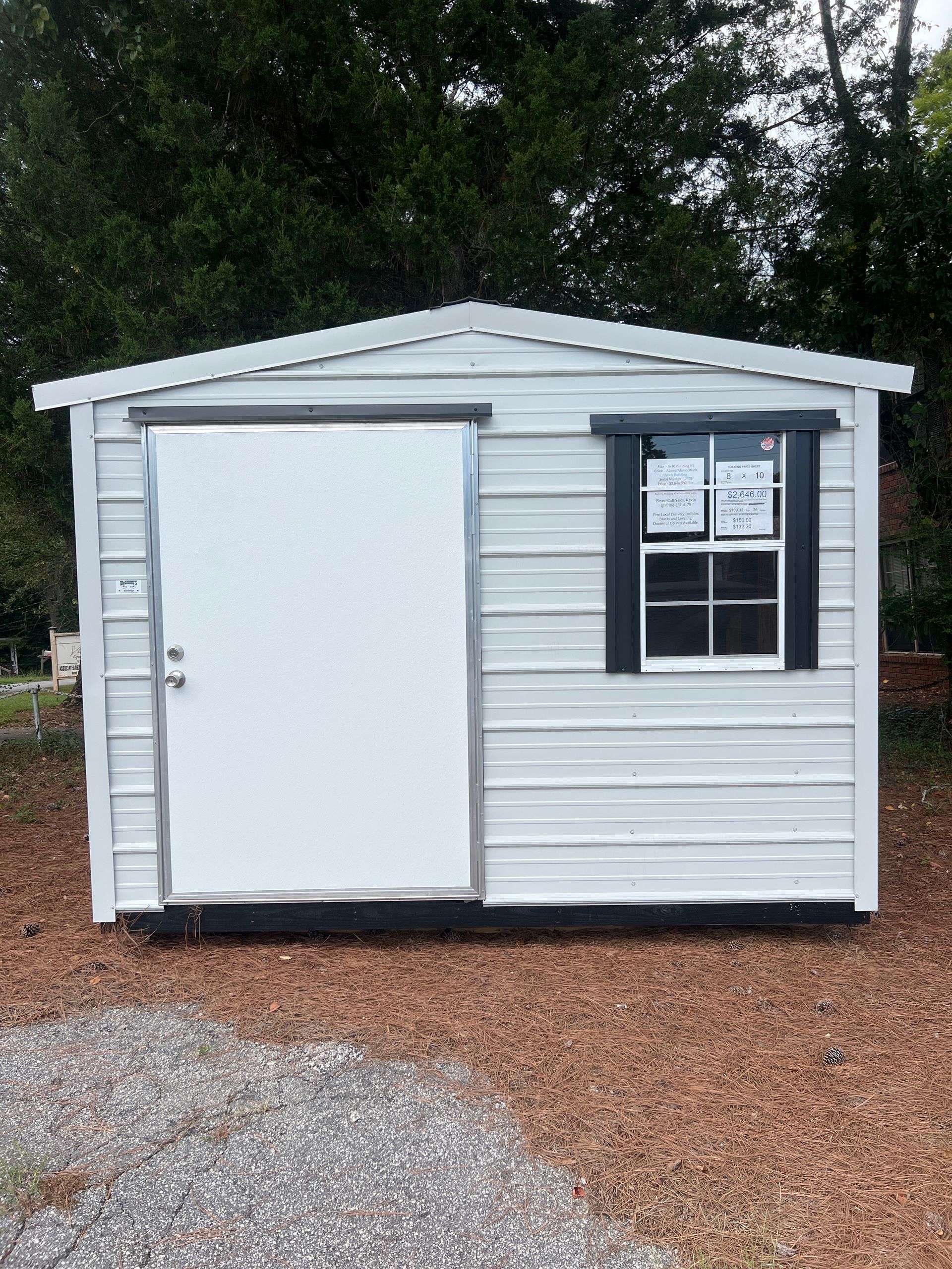 White shed with a sliding door, a black-framed window, on a gravel and pine straw surface.