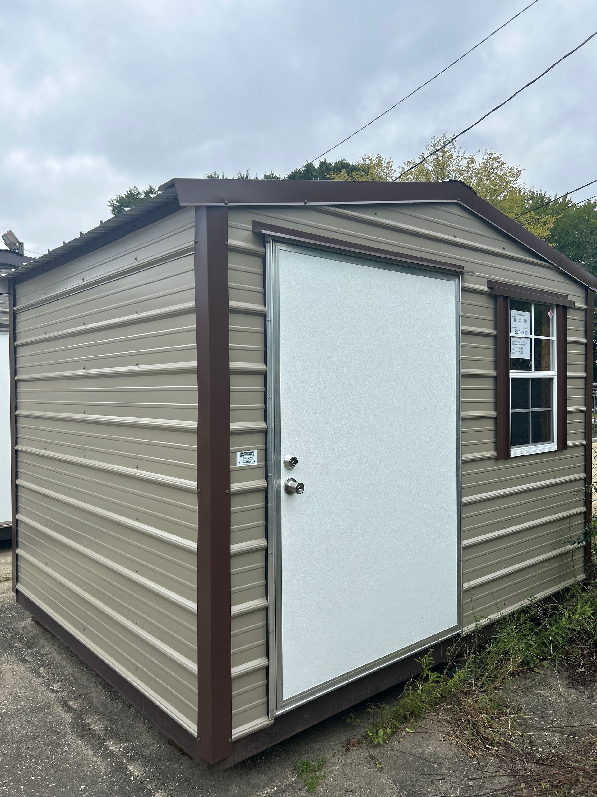 Tan and brown metal storage shed with white door and window. Set outdoors on a cloudy day.