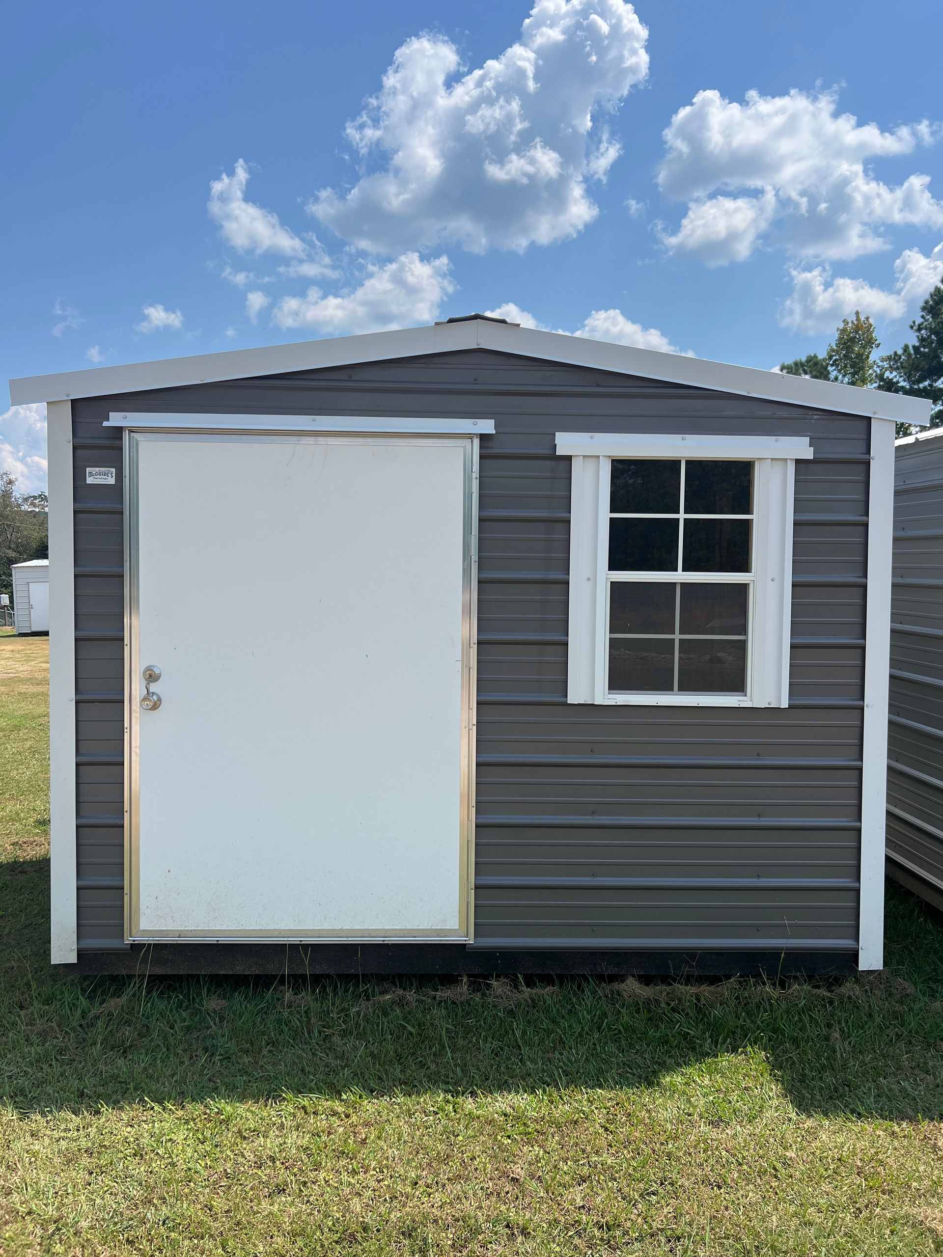 Gray metal shed with white door, window, and trim, on grass under a blue sky with clouds.