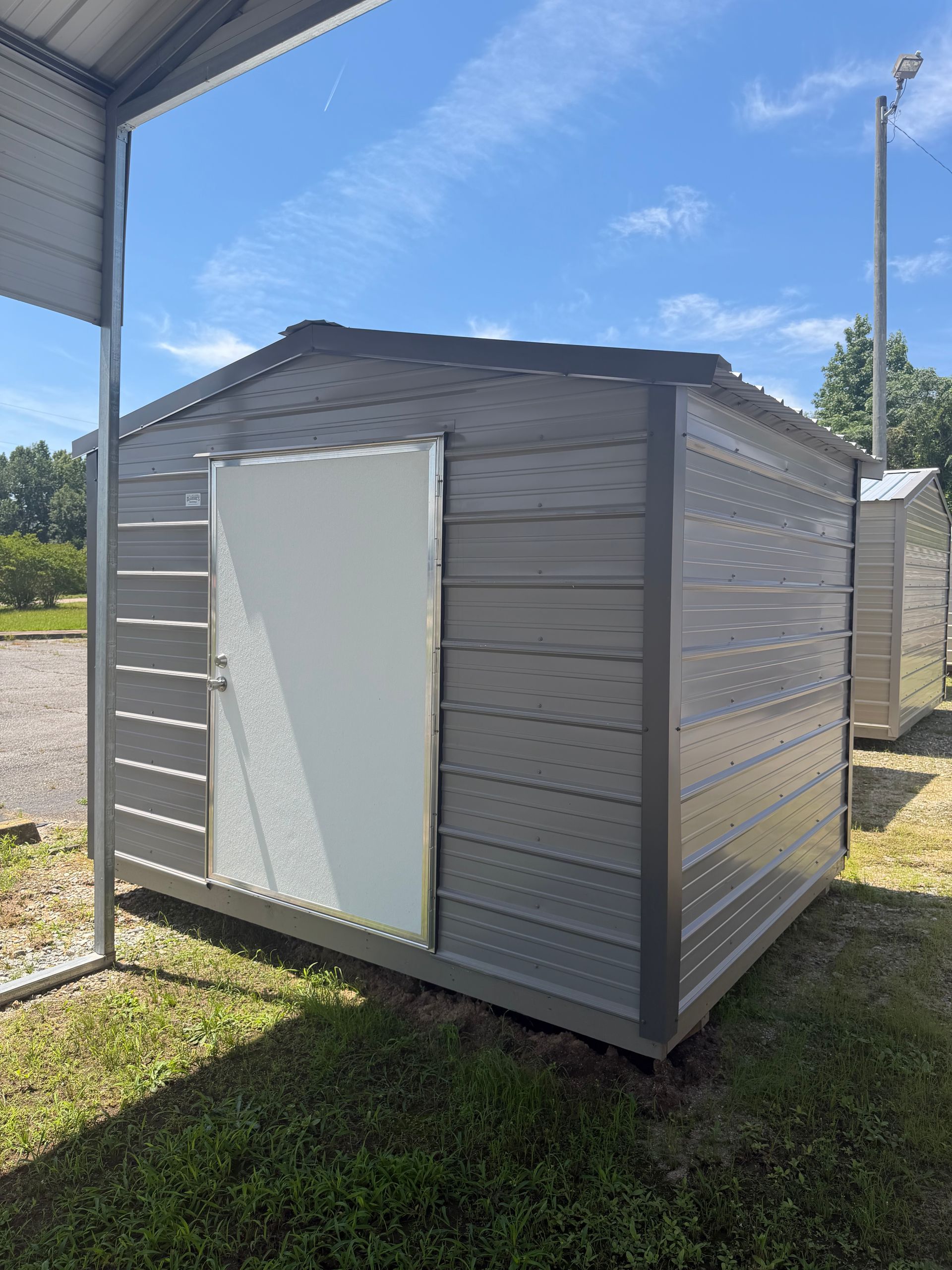 Small gray metal storage shed with a white door, standing outdoors on grass in bright sunlight.