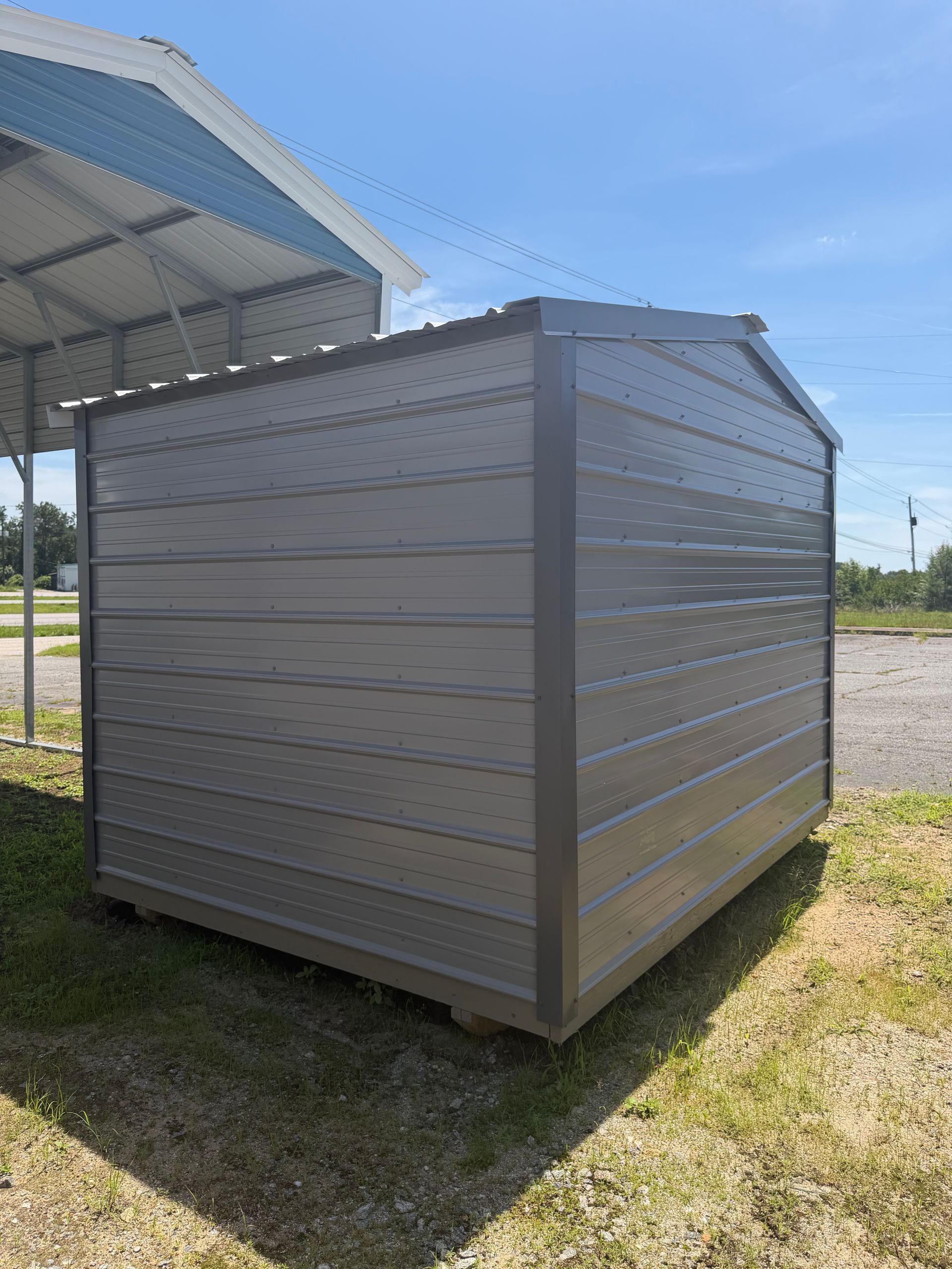 Gray metal storage shed beside a covered structure on a grassy lot under a blue sky