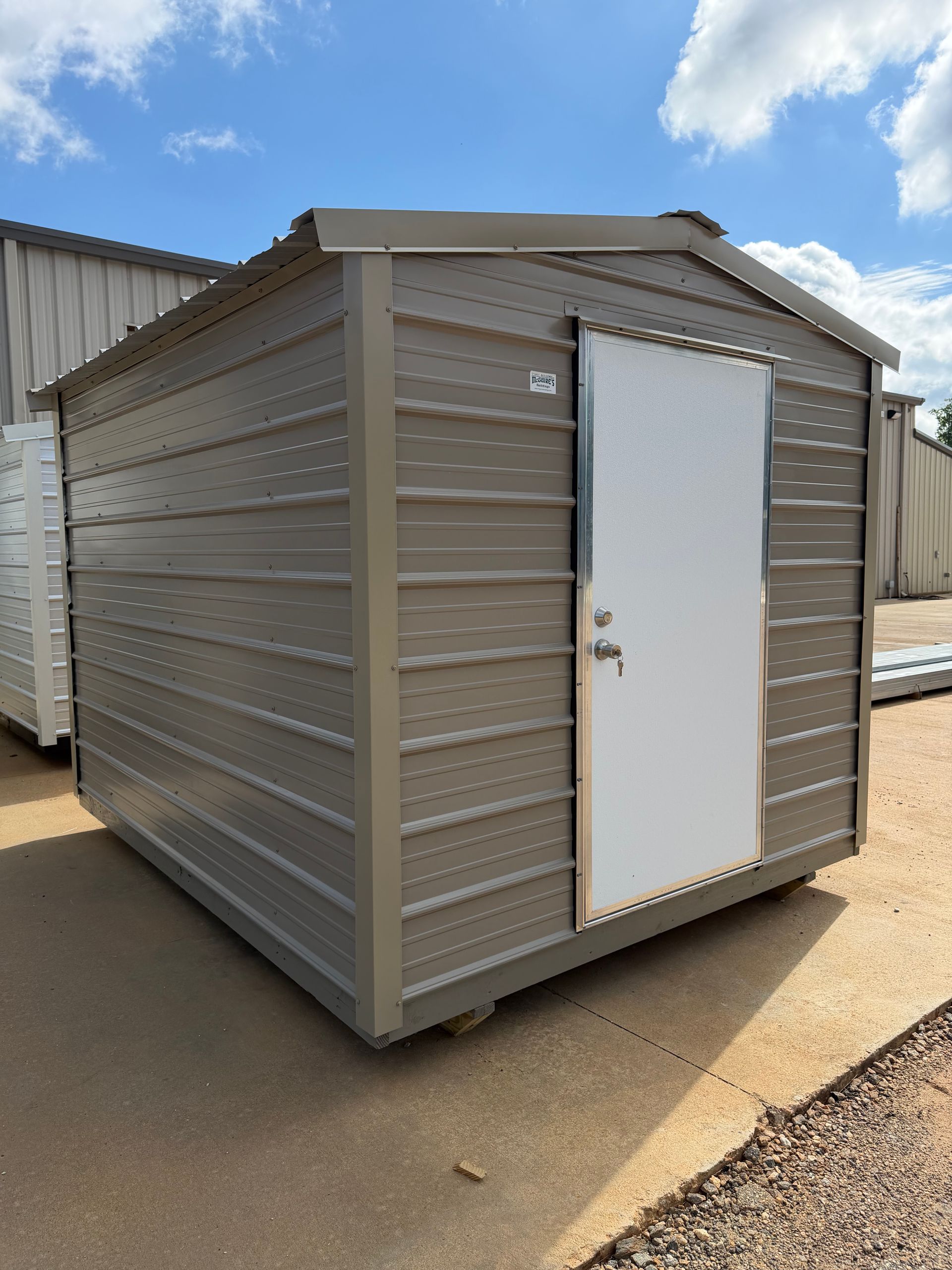 Small gray metal storage shed with a white door on a paved lot.