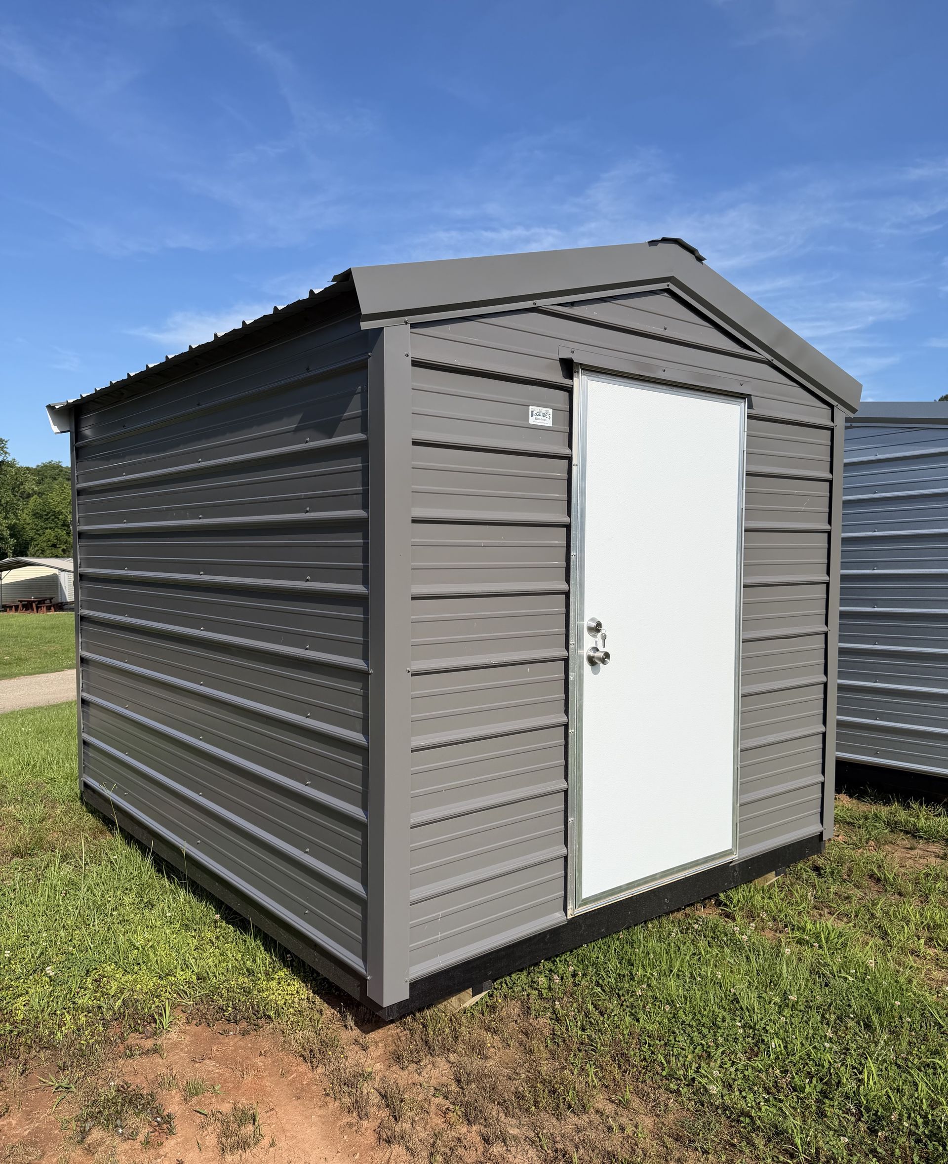 Gray storage shed with white door on a grassy lot under a blue sky